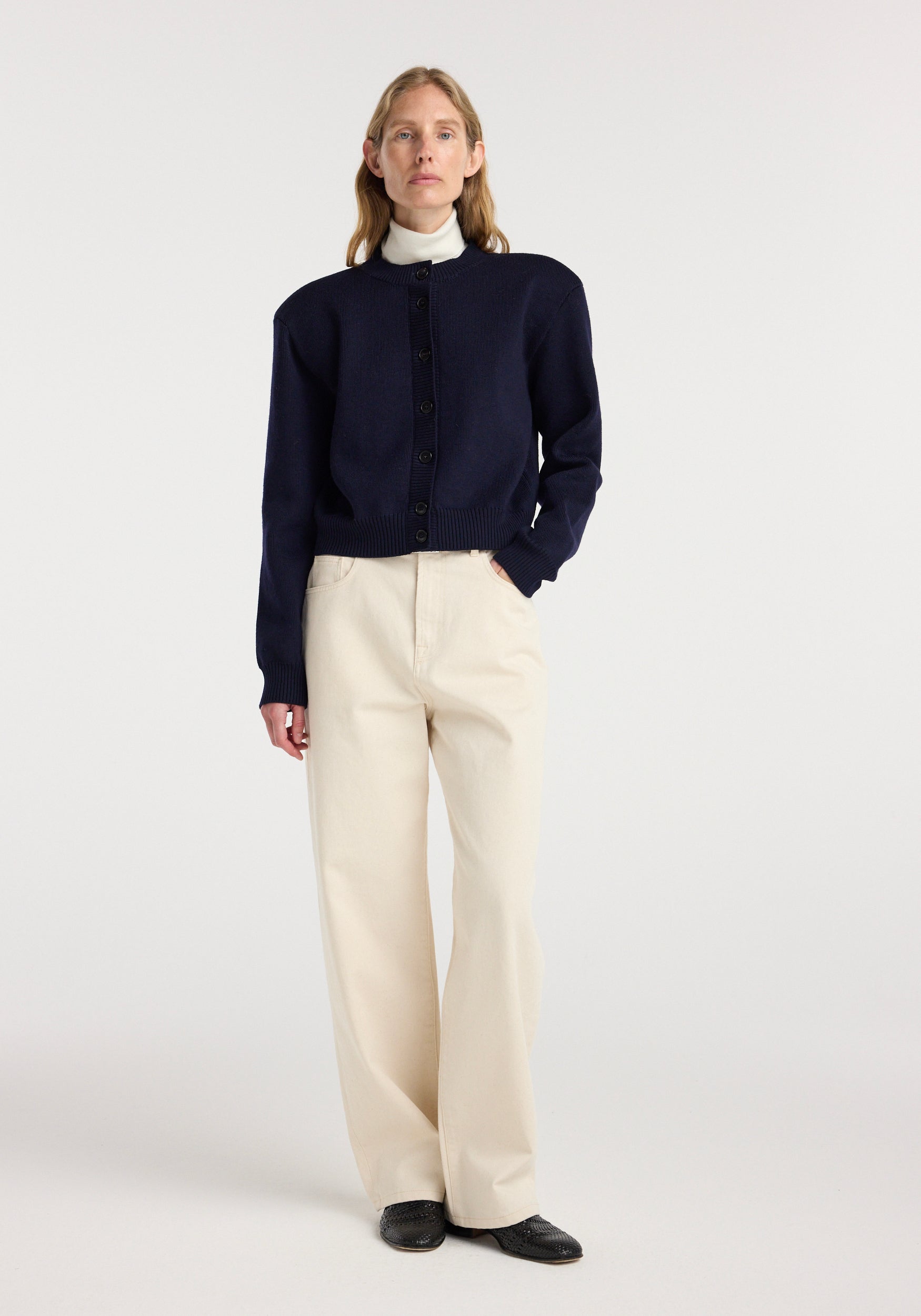 Woman wearing a navy button-up Statement shoulder cardigan | navy with a white turtleneck underneath, paired with beige wide-leg pants and black shoes, standing against a plain background.