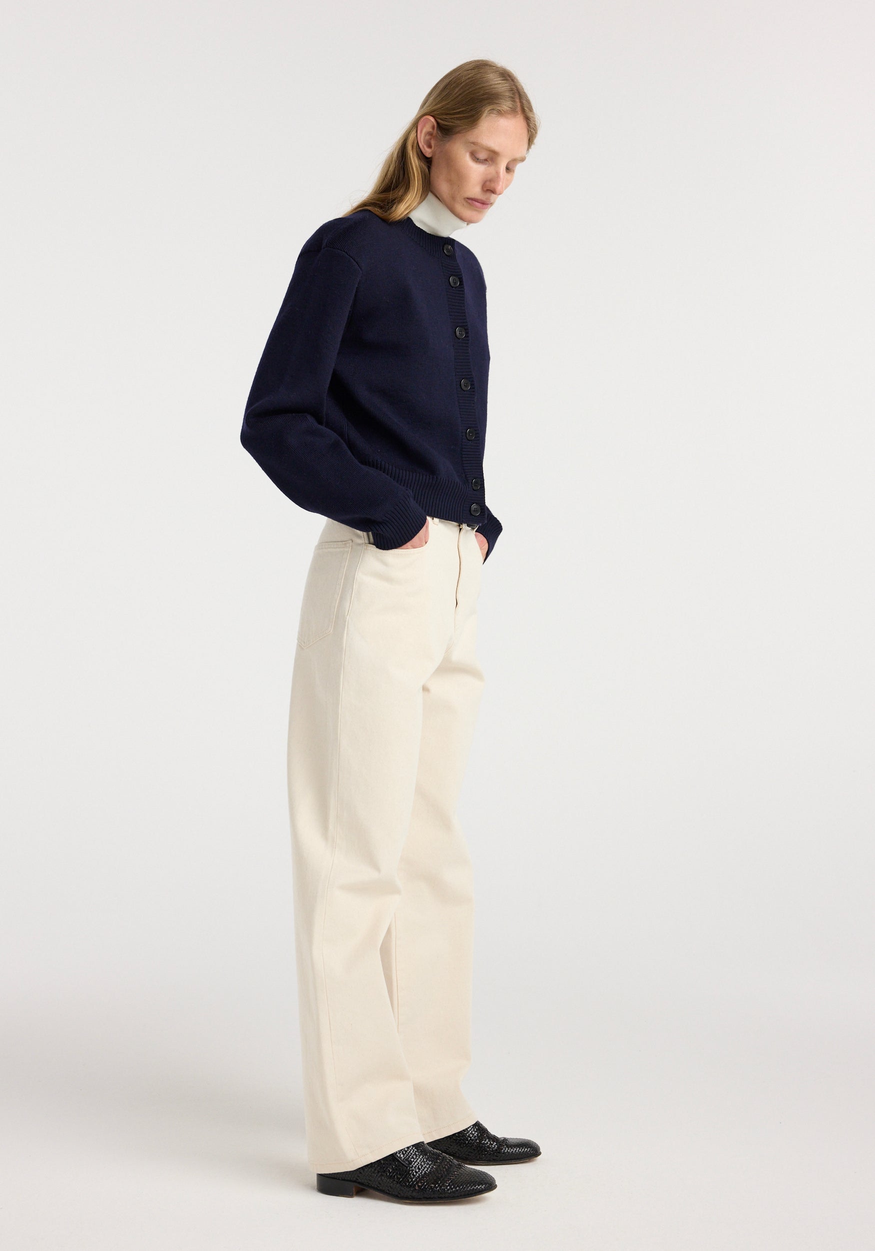 Model wearing a navy Statement shoulder cardigan | navy with long sleeves and black buttons, paired with cream wide-leg pants and black textured shoes, standing against a white background.