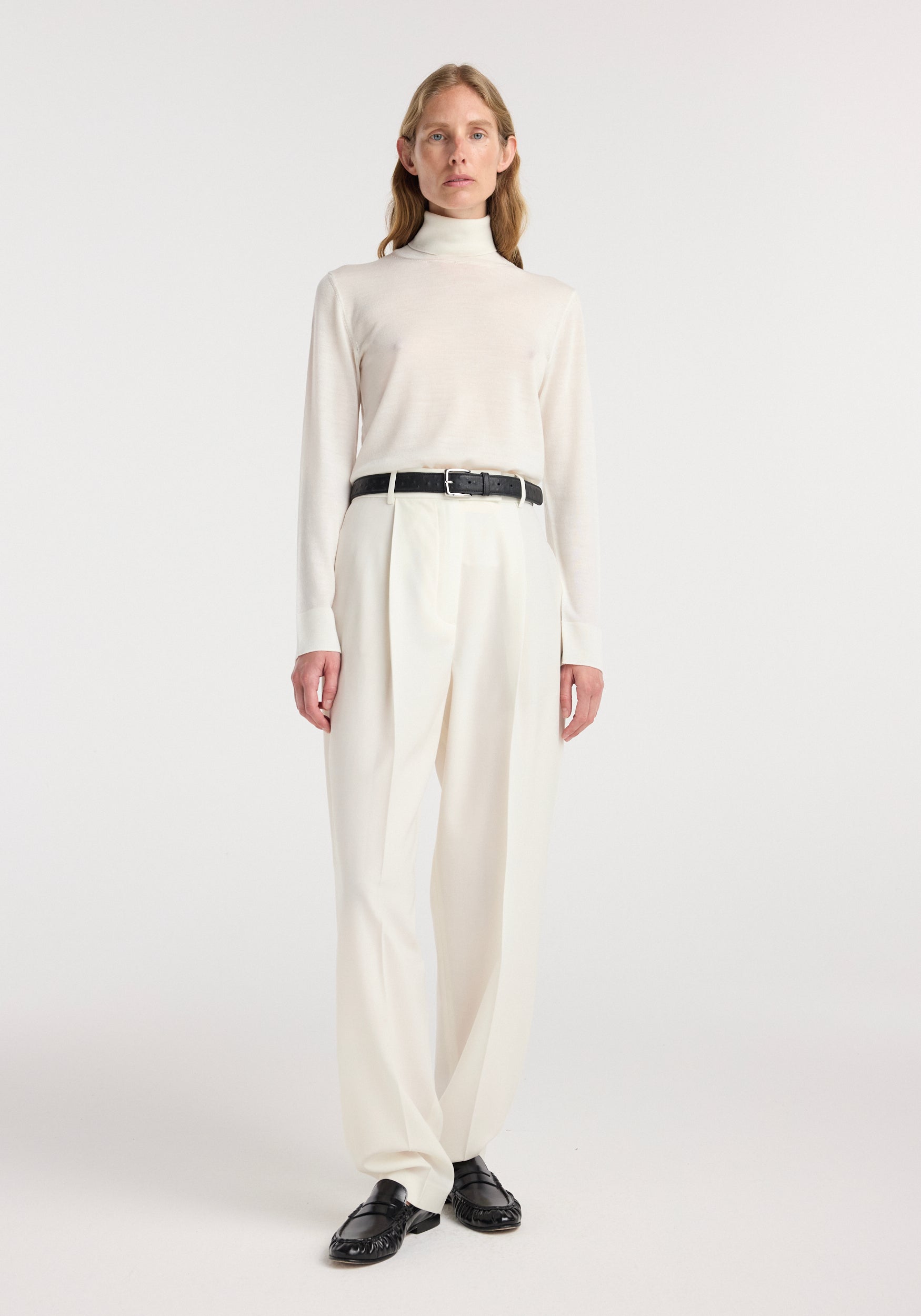 Model wearing a Fine-knit wool cashmere turtleneck | off white paired with high-waisted cream trousers, a black belt, and black loafers, standing against a plain white background.