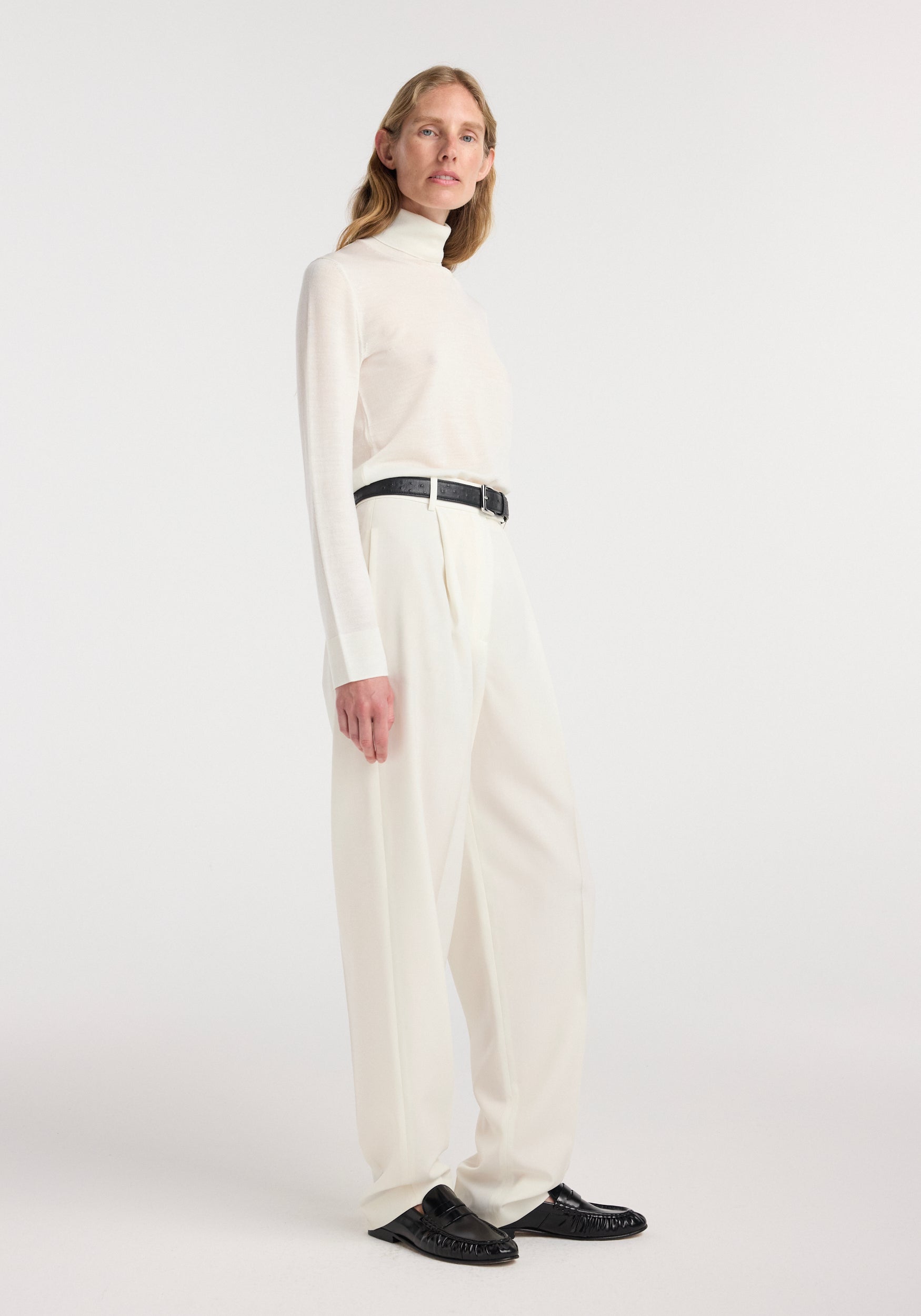 Model wearing a Fine-knit wool cashmere turtleneck | off white paired with matching high-waisted trousers and black belt, standing against a plain light background.