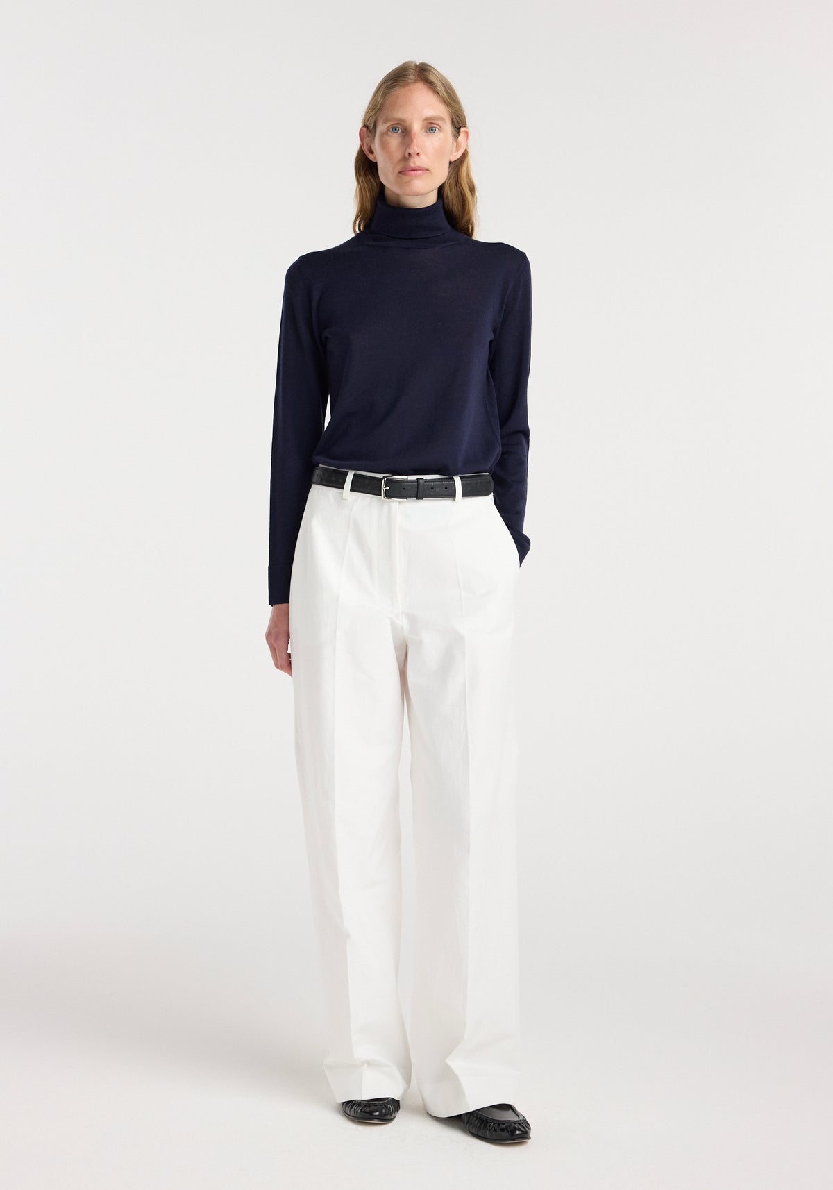 Woman wearing a navy fine-knit wool cashmere turtleneck paired with white wide-leg trousers, black belt, and black shoes, standing against a plain white background.