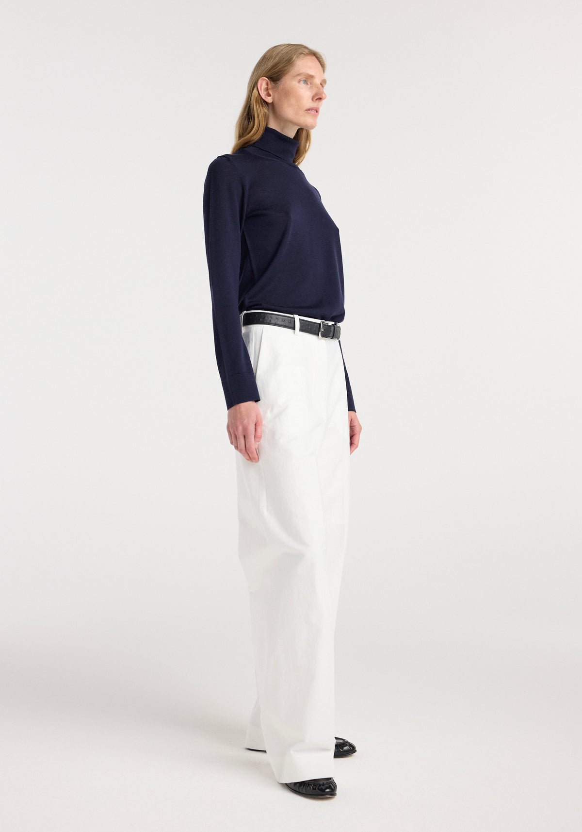 Woman wearing a navy fine-knit wool cashmere turtleneck paired with high-waisted white wide-leg trousers and a black belt, standing against a plain background.