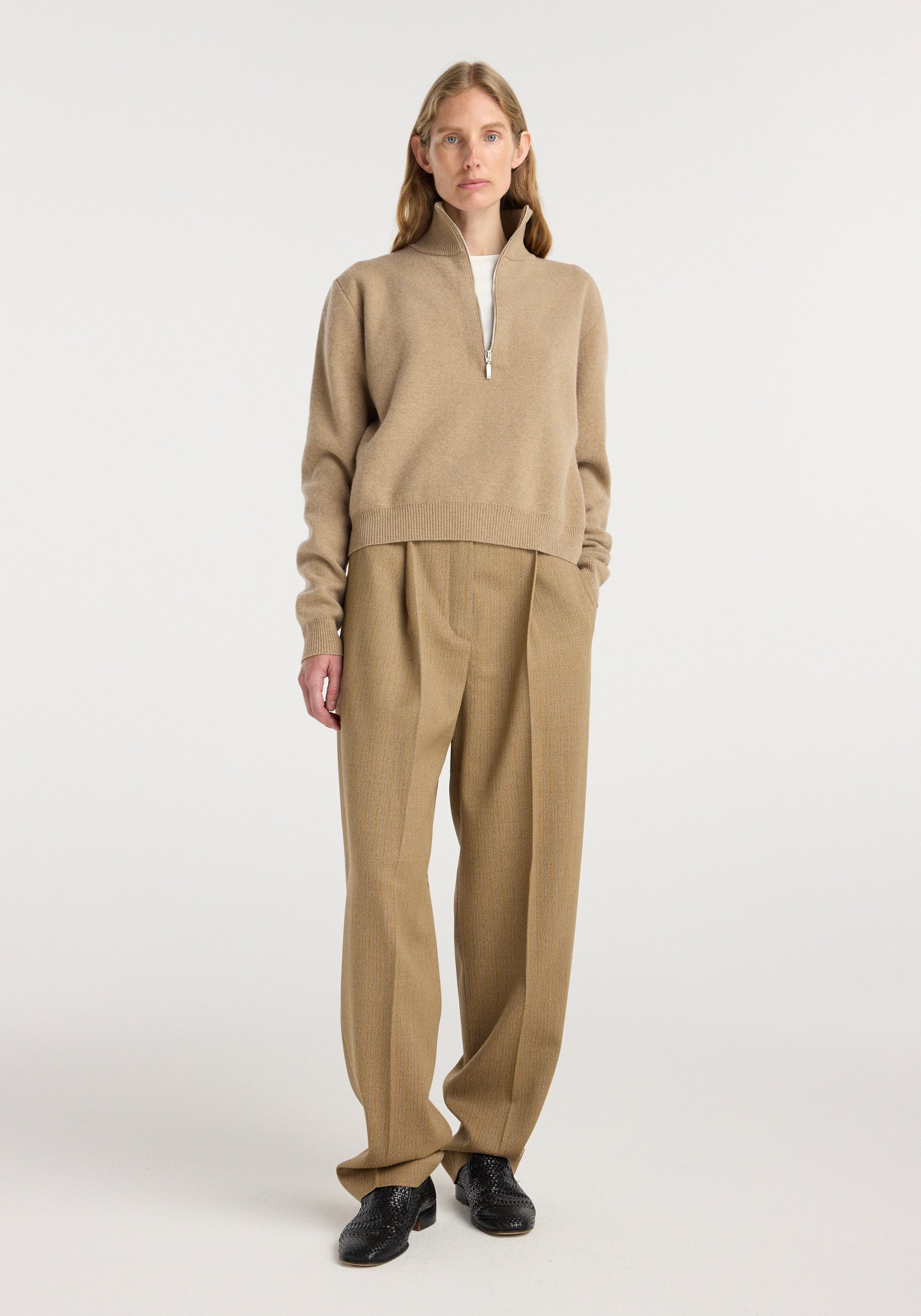 Woman wearing beige Daria - Single pleat tapered trousers | heritage stripe paired with a light brown zip-up sweater and black shoes, standing against a plain white background.