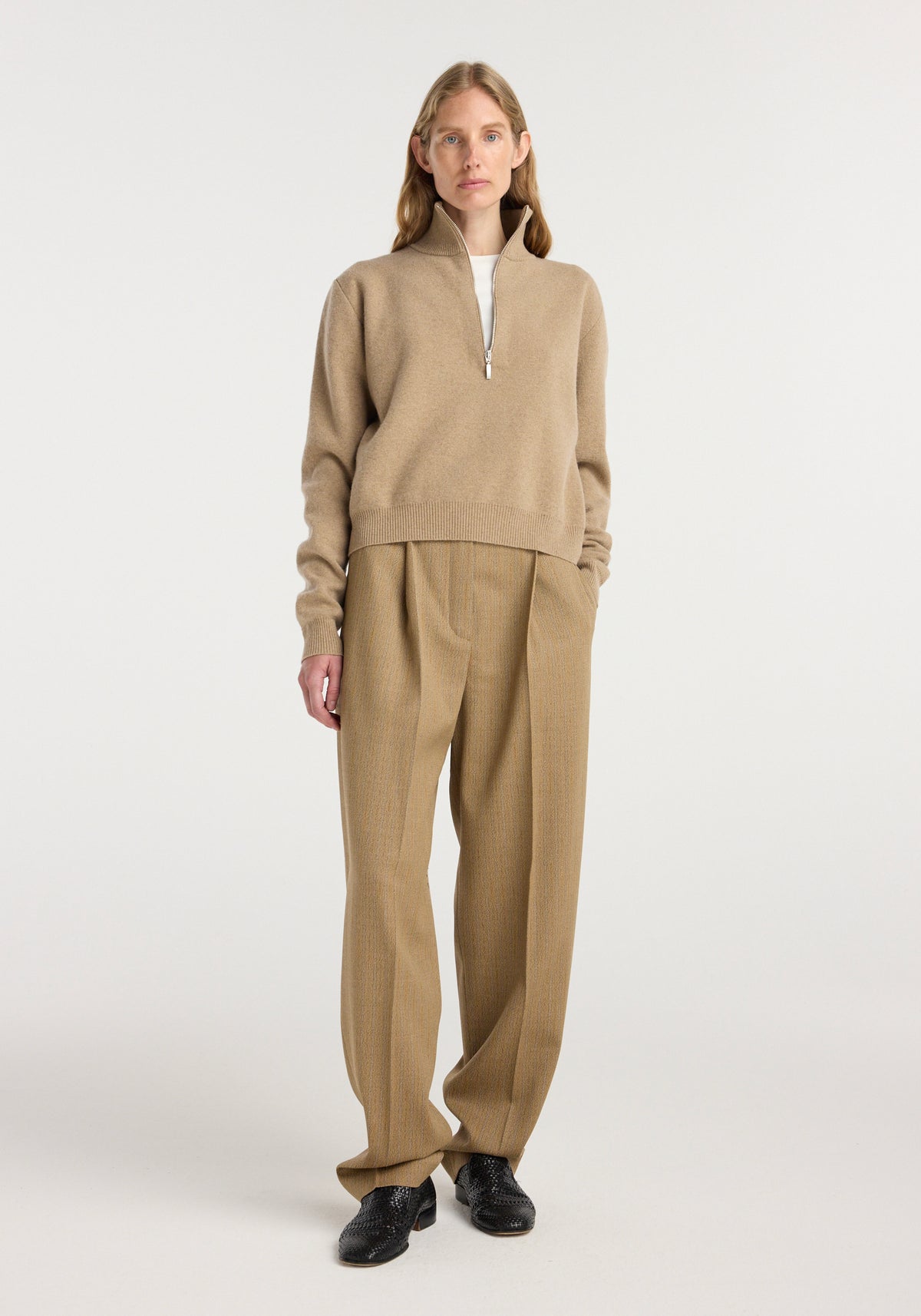 A woman wearing a zipped mouliné polo in camel color with long sleeves and a high collar, paired with loose camel trousers and black shoes, standing against a plain background.