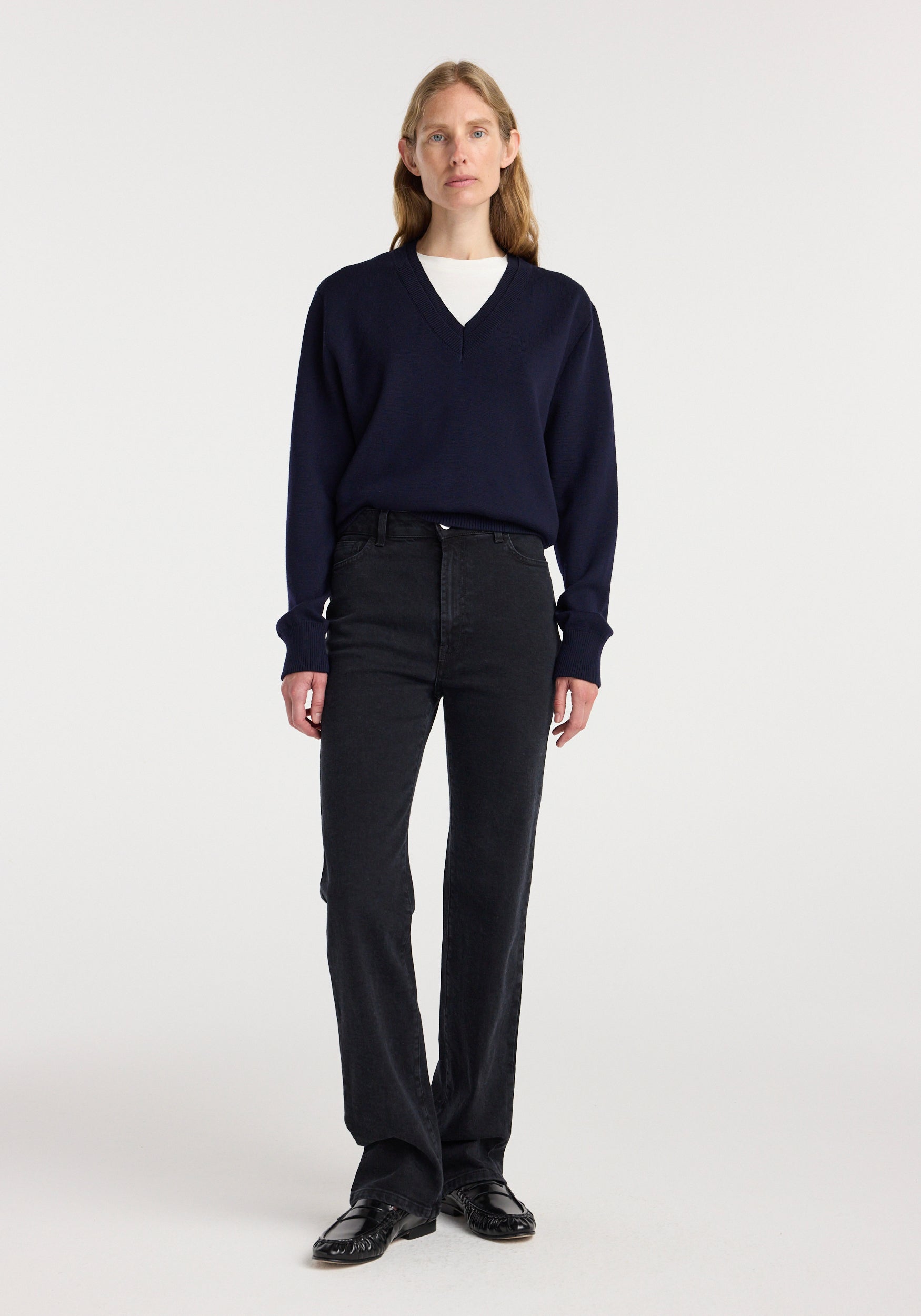 A woman wearing Lino - high rise straight denim | black paired with a dark navy v-neck sweater and black shoes, standing against a plain white background.