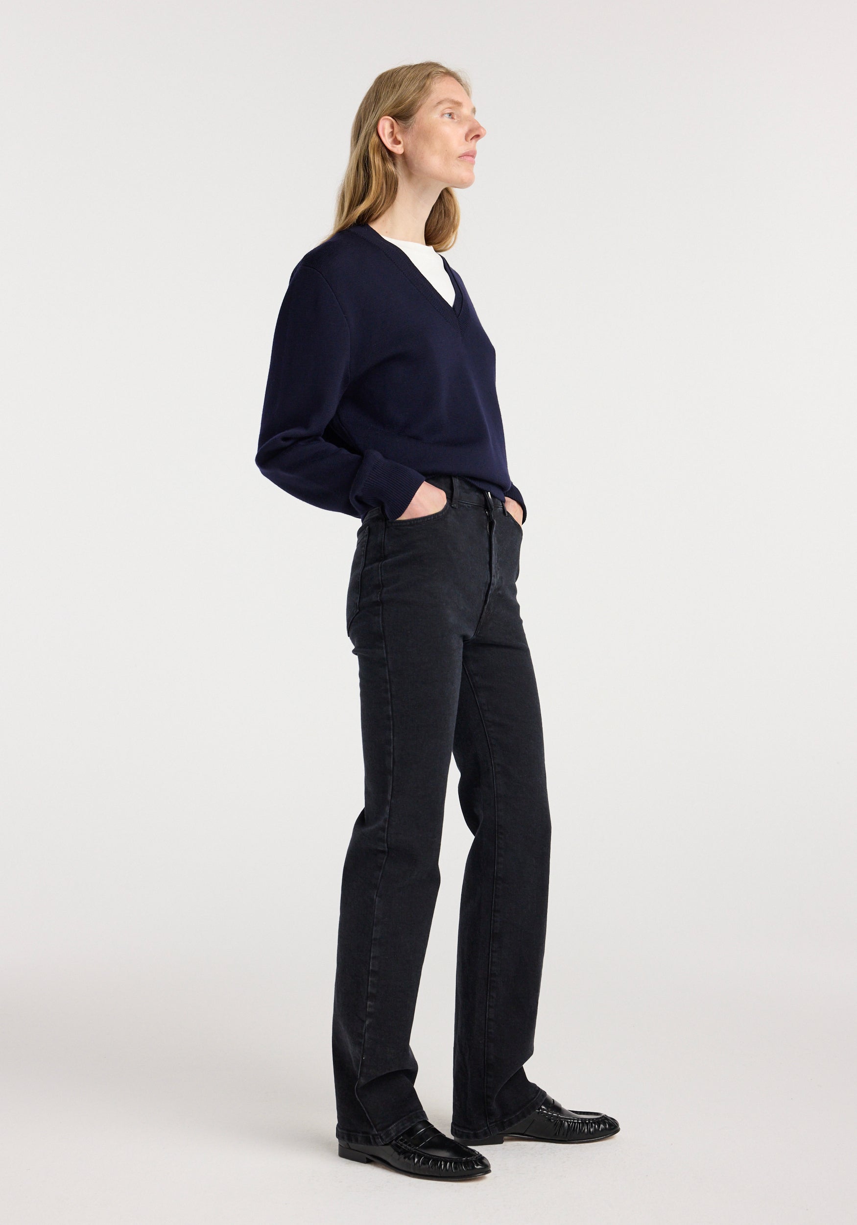 Woman wearing Lino - high rise straight denim | black jeans with hands in pockets, paired with a dark navy sweater and black loafers, standing against a plain light background.