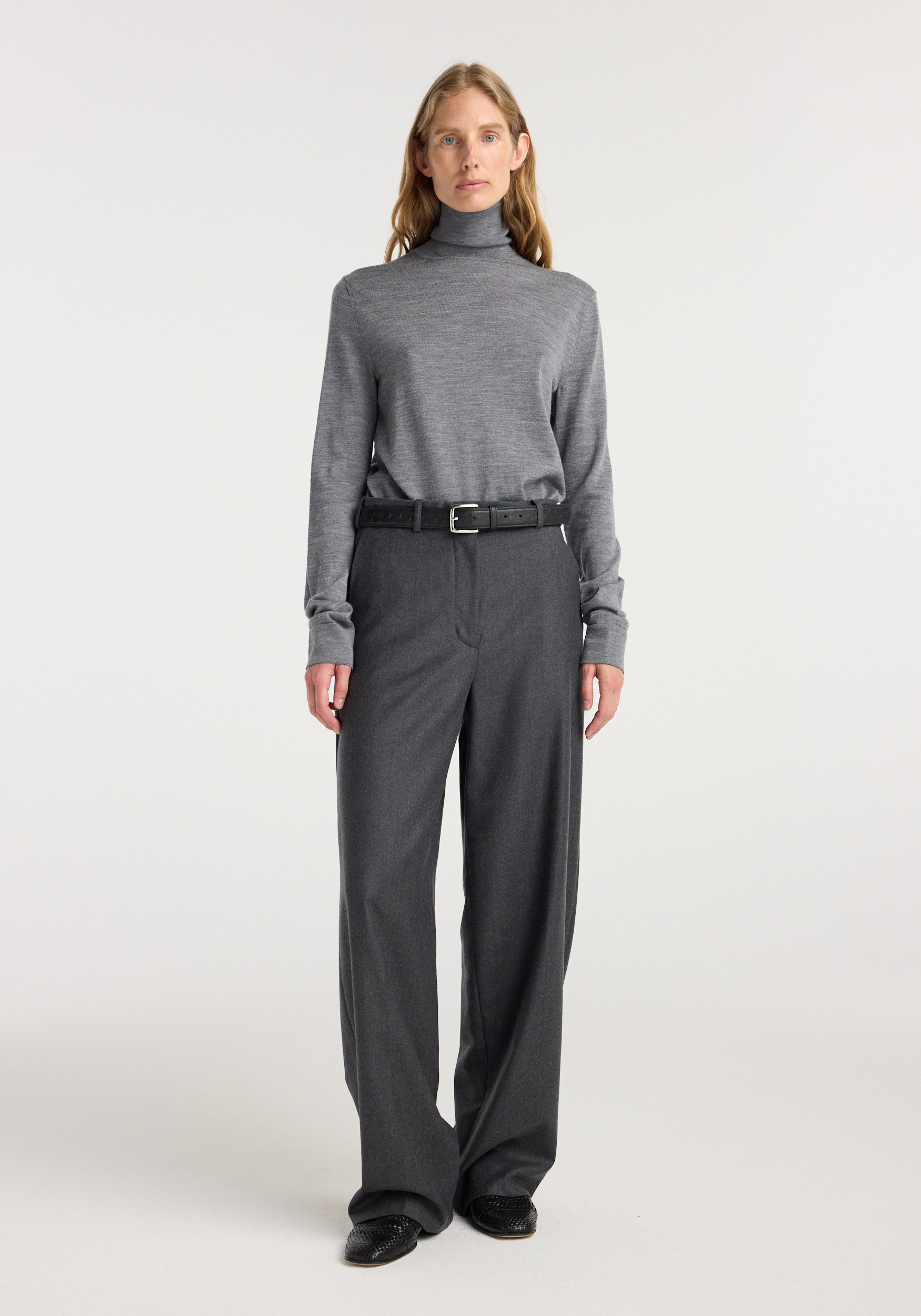 A woman wearing a fine knitted wool turtleneck | mid grey melange paired with high-waisted dark grey wide-leg trousers and black shoes, standing against a plain light background.