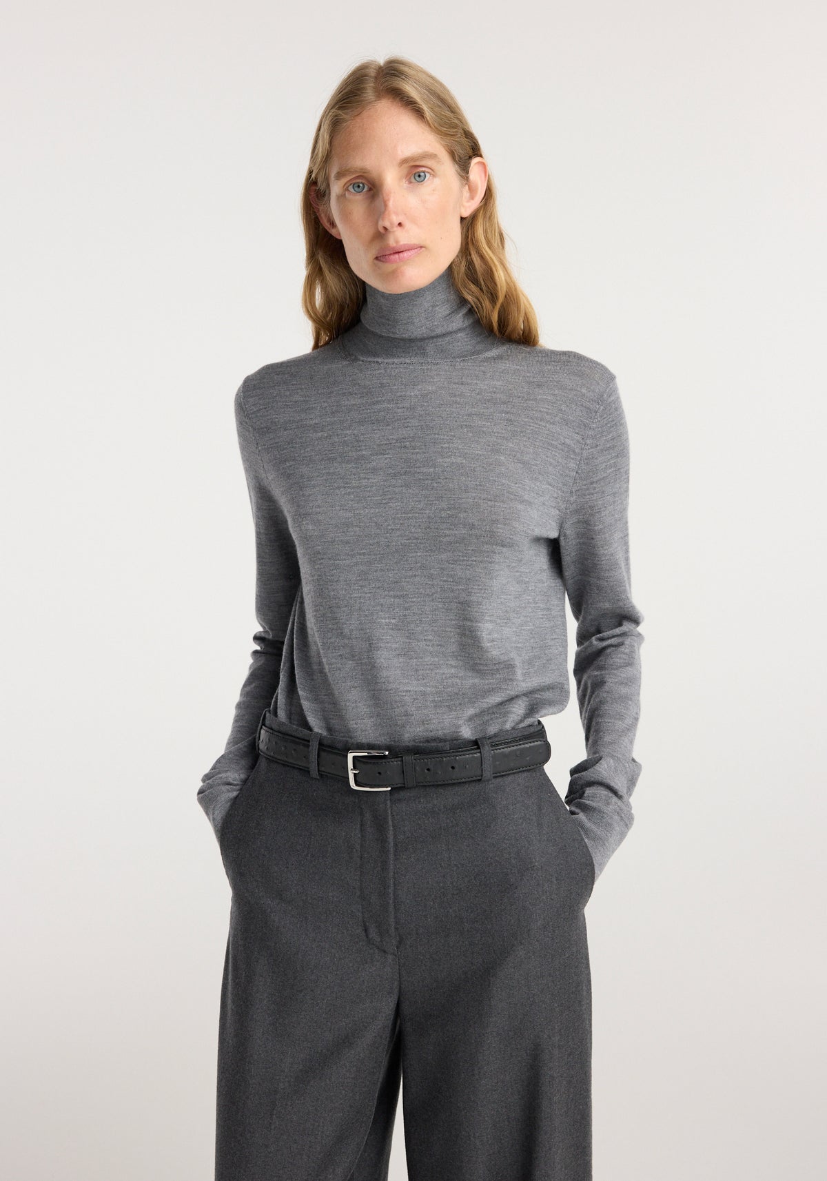 Woman wearing a fine knitted wool turtleneck | mid grey melange paired with dark grey high-waisted trousers and a black belt, standing against a plain light background.