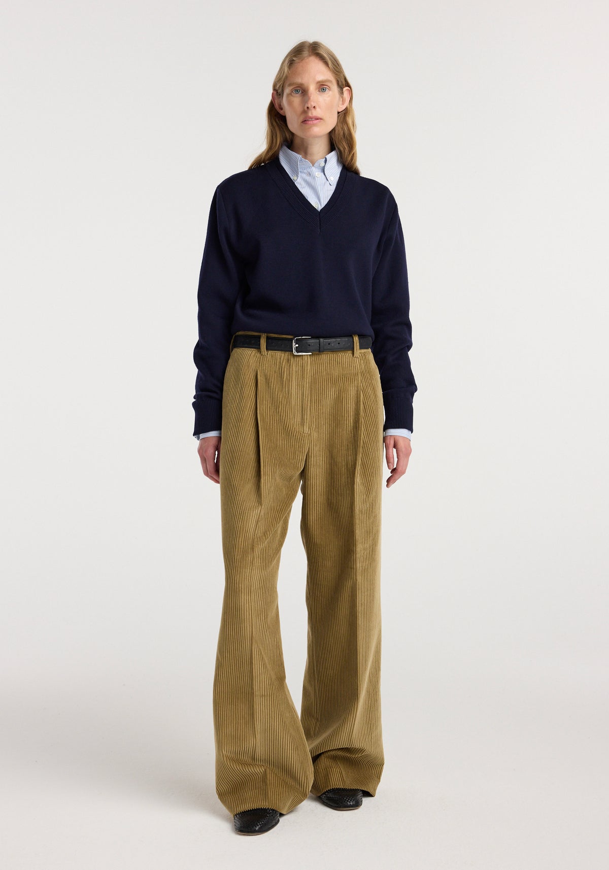 Woman wearing Ted - Lower waist corduroy trousers | beige with pleats, styled with a black belt, navy V-neck sweater, light blue button-up shirt, and black shoes against a neutral background.