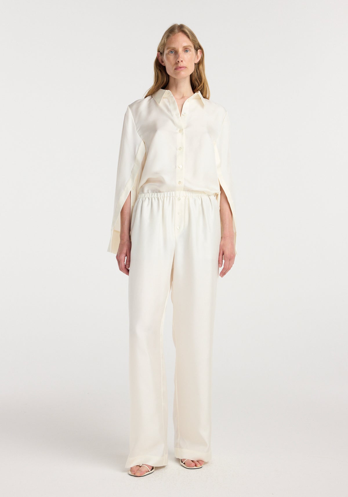 Woman wearing cream-colored silk relaxed trousers with an elastic waistband and a matching silk button-up shirt, standing against a plain white background.