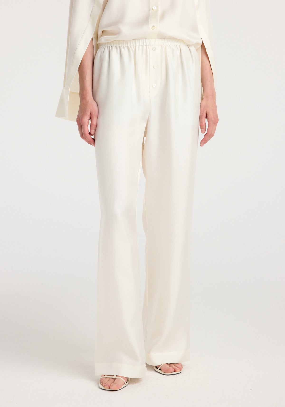 Front view of a person wearing cream silk relaxed trousers with an elastic waistband, paired with a matching light cream button-up top and white open-toe sandals.