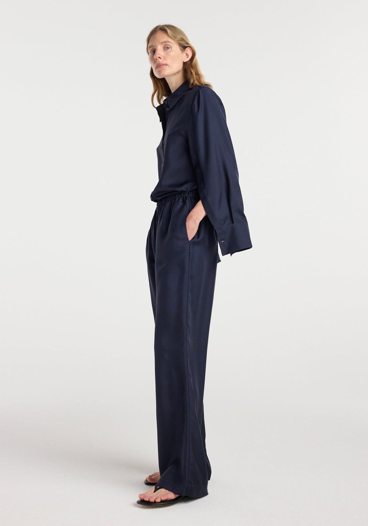 Woman wearing dark navy silk relaxed trousers | night with a matching long-sleeve shirt, standing in a side profile pose against a plain light background.