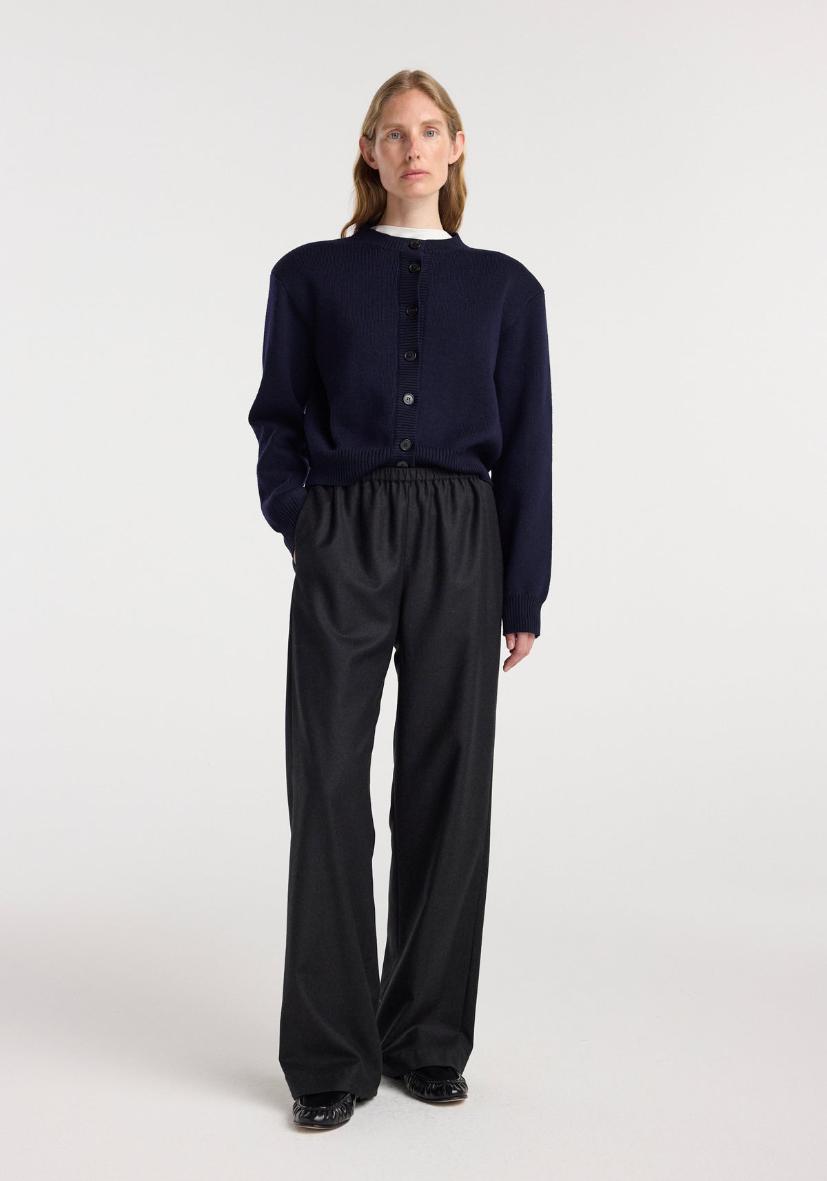 A model wearing relaxed fit trousers | anthracite melange, styled with a navy cardigan and black shoes, standing against a plain white background.