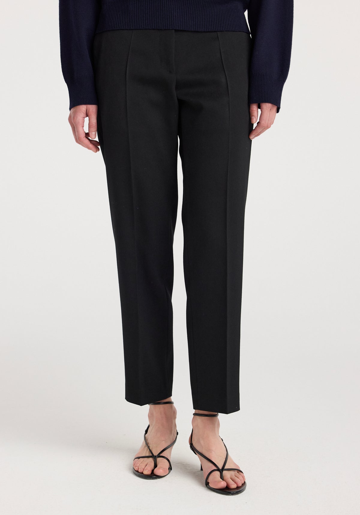 Front view of a person wearing Mies - Cigarette leg tailored trousers | black with a navy sweater and black strappy sandals on a white background