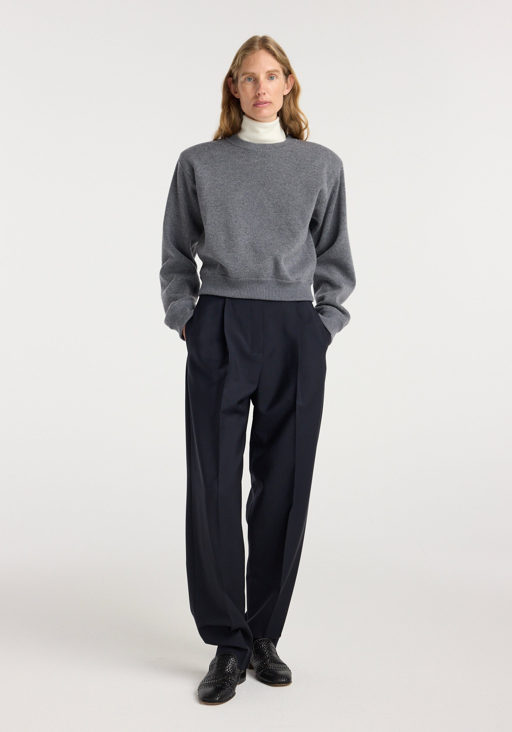 Woman wearing a grey sweatshirt and white turtleneck paired with navy tailored Daria - Single pleat tapered trousers | navy and black dress shoes standing against a plain light background.