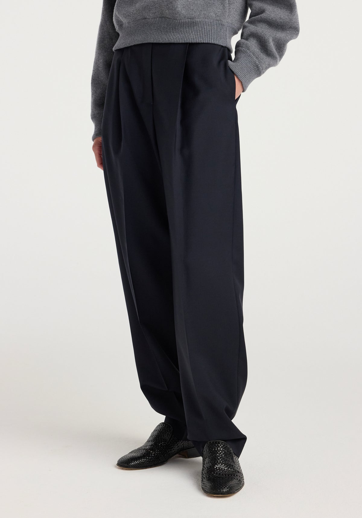 Model wearing Daria - Single pleat tapered trousers | navy paired with a grey sweater and black textured shoes, standing against a plain white background.
