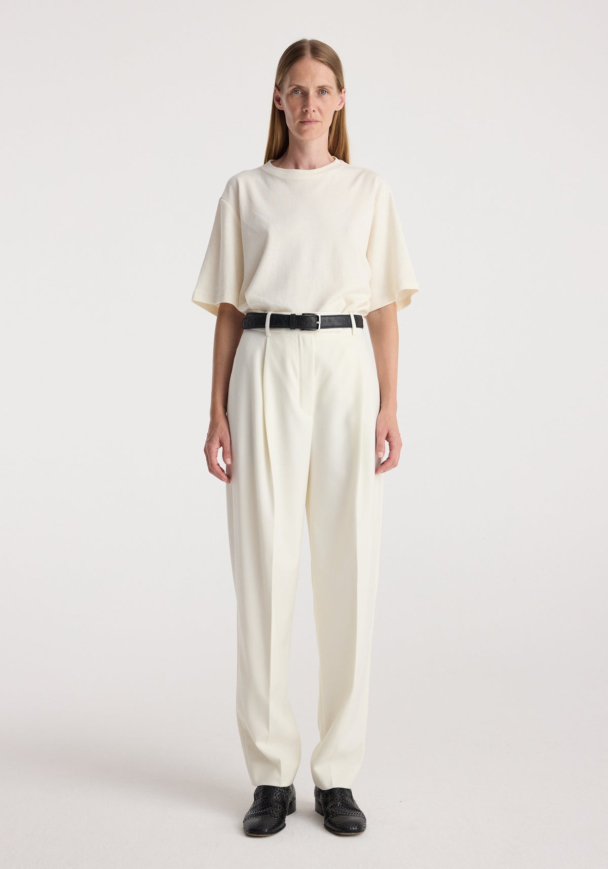 Woman wearing a simple off-white loose-fit short sleeve top tucked into Daria - Single pleat tapered trousers | off white, paired with a black belt and black shoes, standing against a plain background.