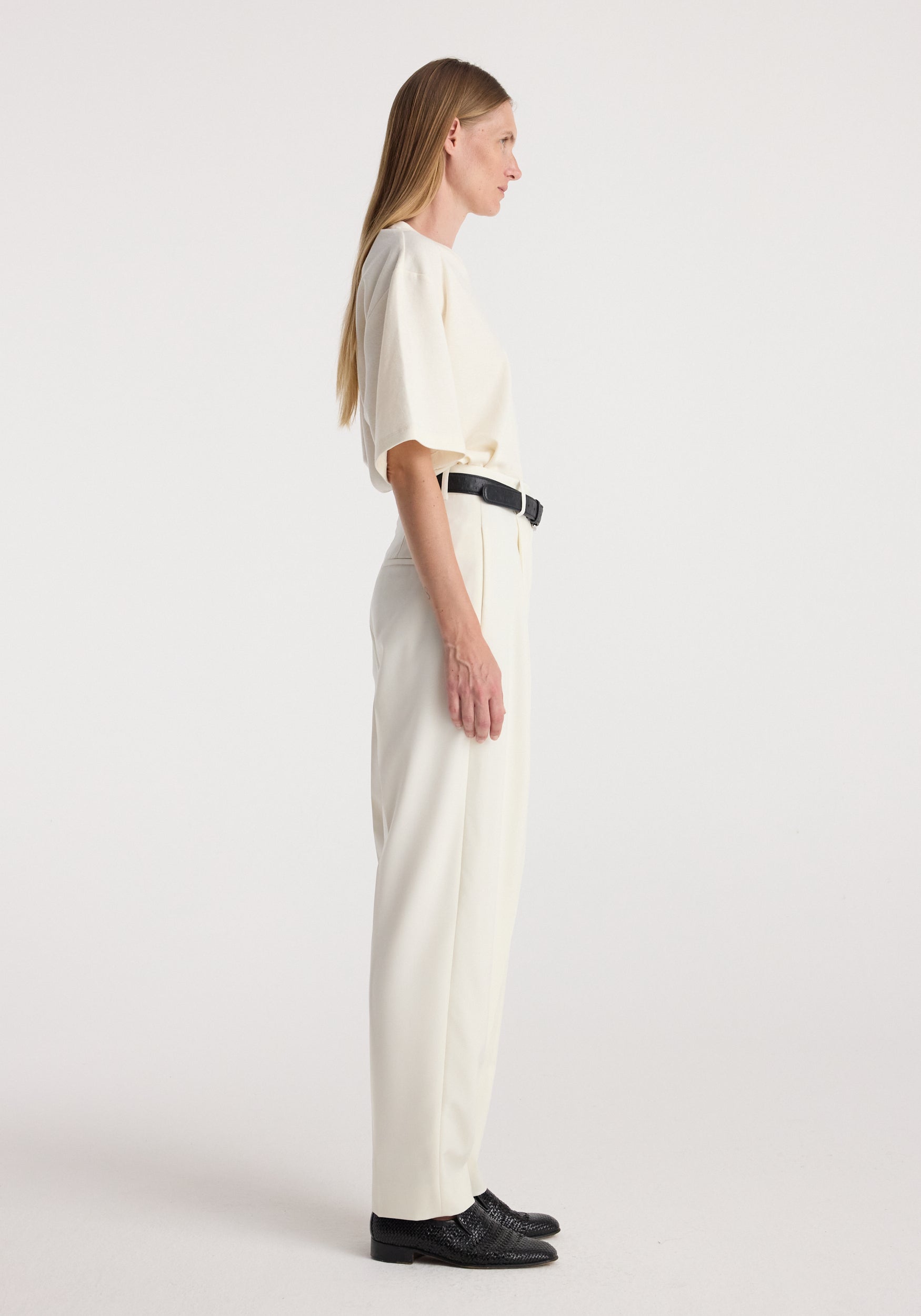 Woman standing in profile wearing Daria - Single pleat tapered trousers | off white paired with a matching light top, black belt, and black shoes against a plain background.