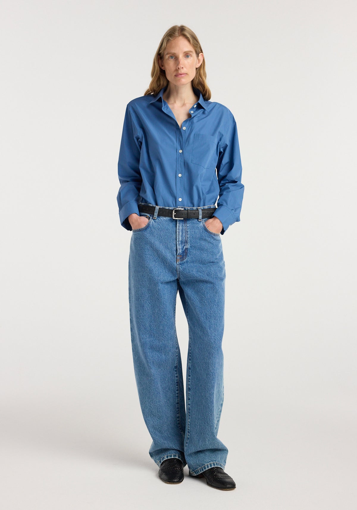 Woman wearing a blue button-up shirt tucked into Bruun - Signature baggy denim | heritage blue jeans with black belt and black shoes, standing against a plain background.