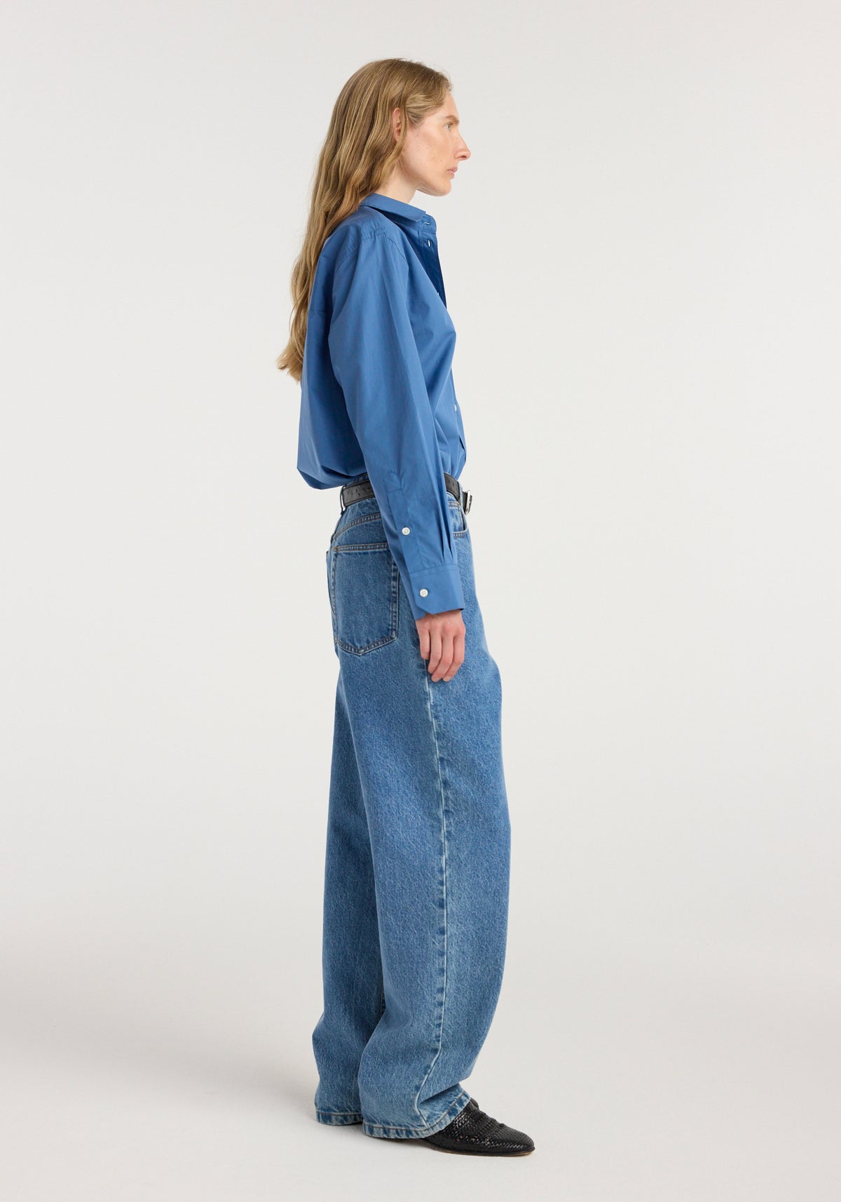 Side view of a woman wearing Bruun - Signature baggy denim | heritage blue paired with a tucked-in blue shirt and black shoes against a plain background.