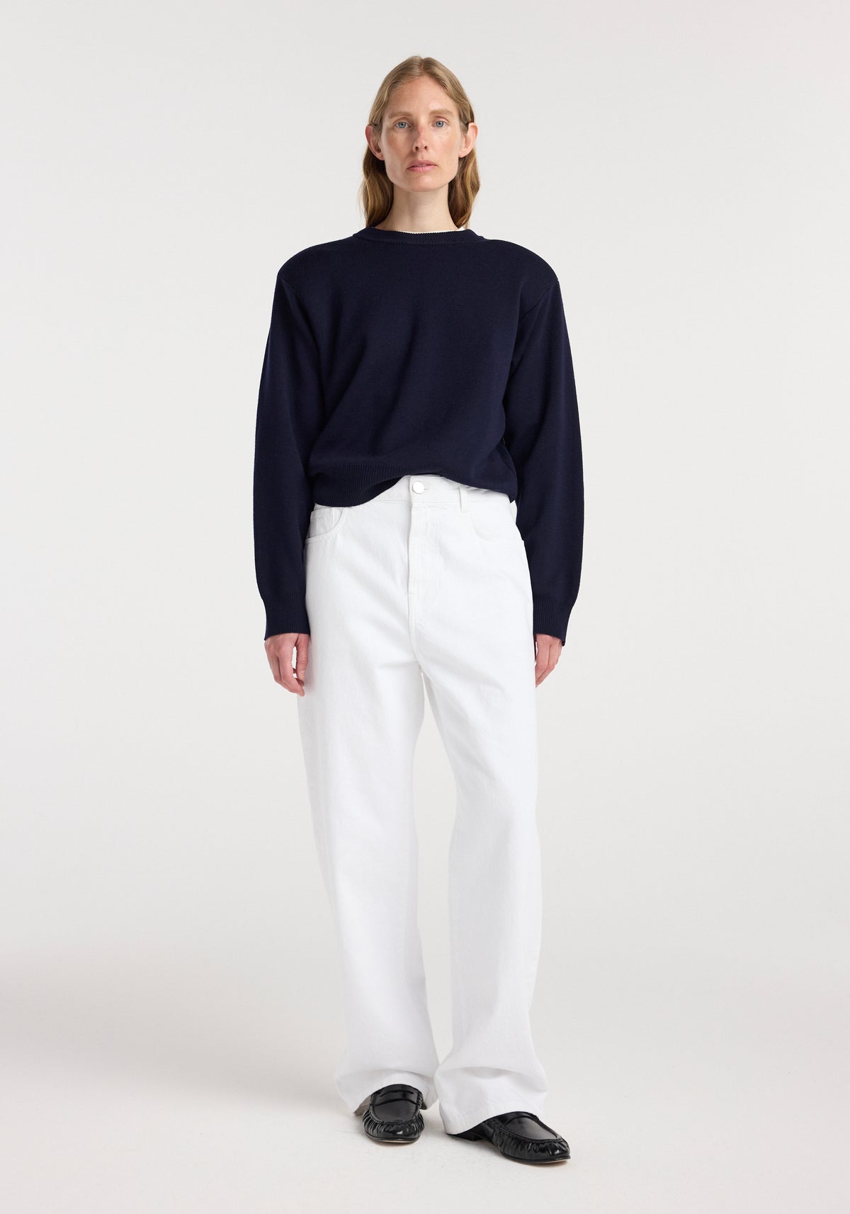 Woman standing wearing a dark navy sweater paired with white loose-fitting pants and black shoes, styled as Bruun - Signature baggy denim | optic white.