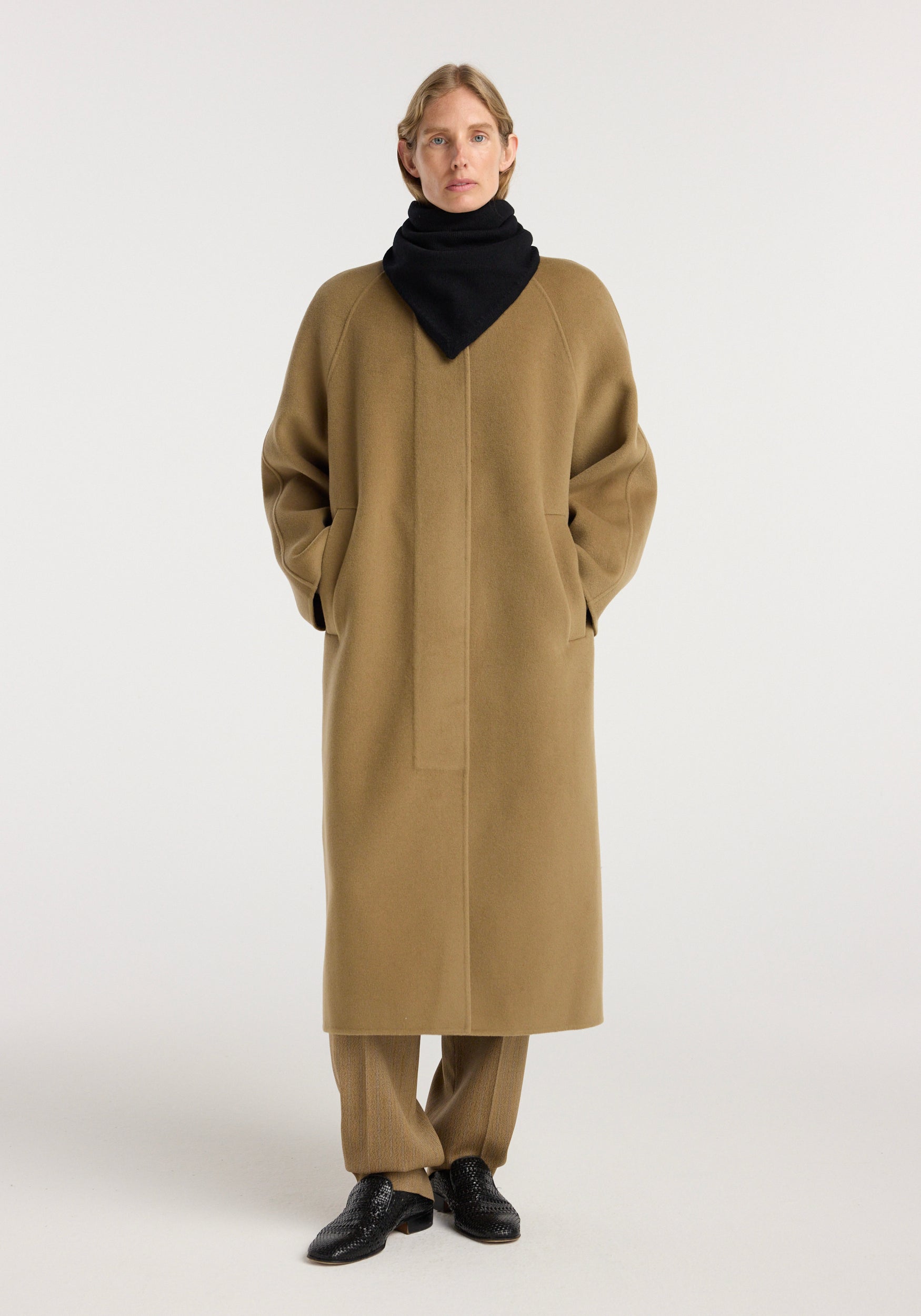 Person wearing a black Small knitted triangle scarf | black wrapped around the neck, paired with a long camel coat, brown pants, and black shoes against a plain background.