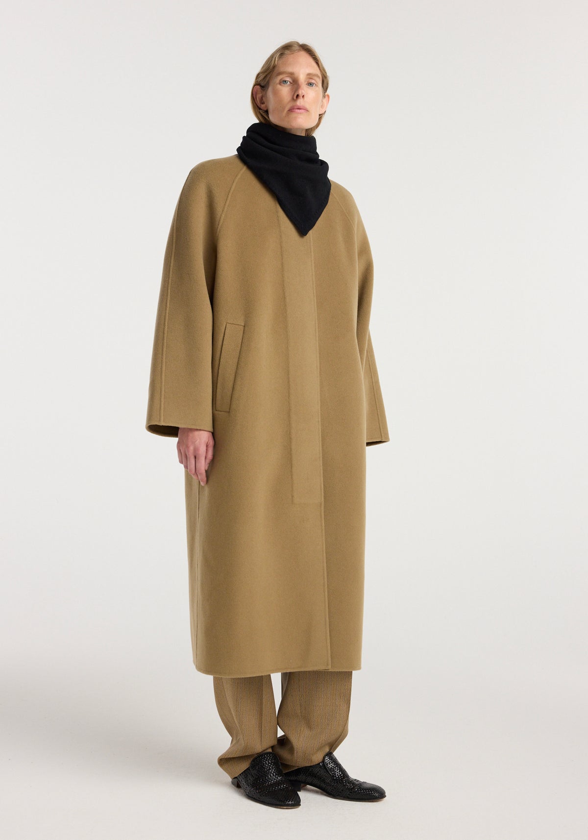 Model wearing a black Small knitted triangle scarf | black styled with a long camel coat, brown pants, and black shoes against a plain white background.