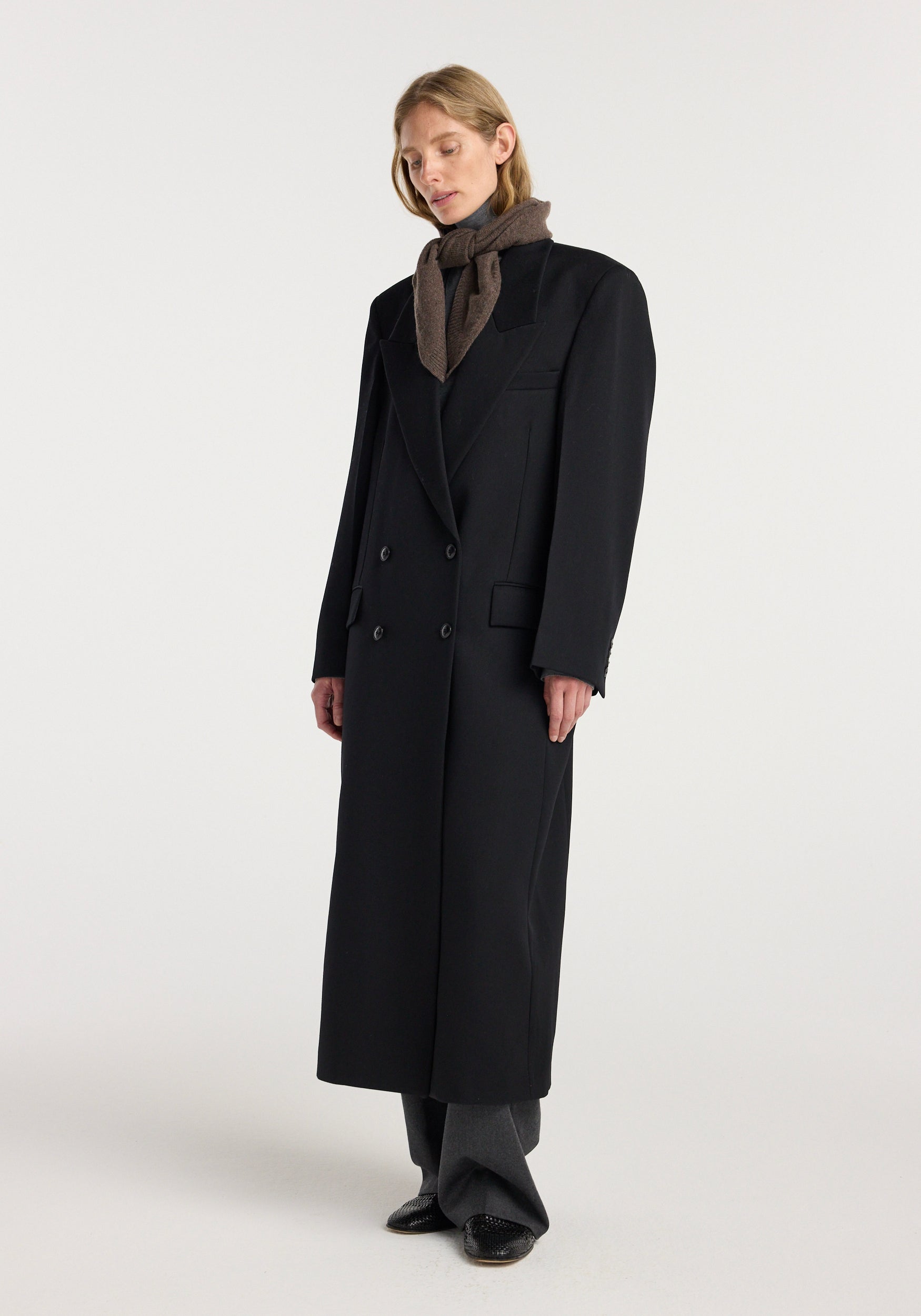 Woman wearing a small knitted triangle scarf | brown melange, styled around the neck with a long black coat, gray trousers, and black shoes against a plain background.