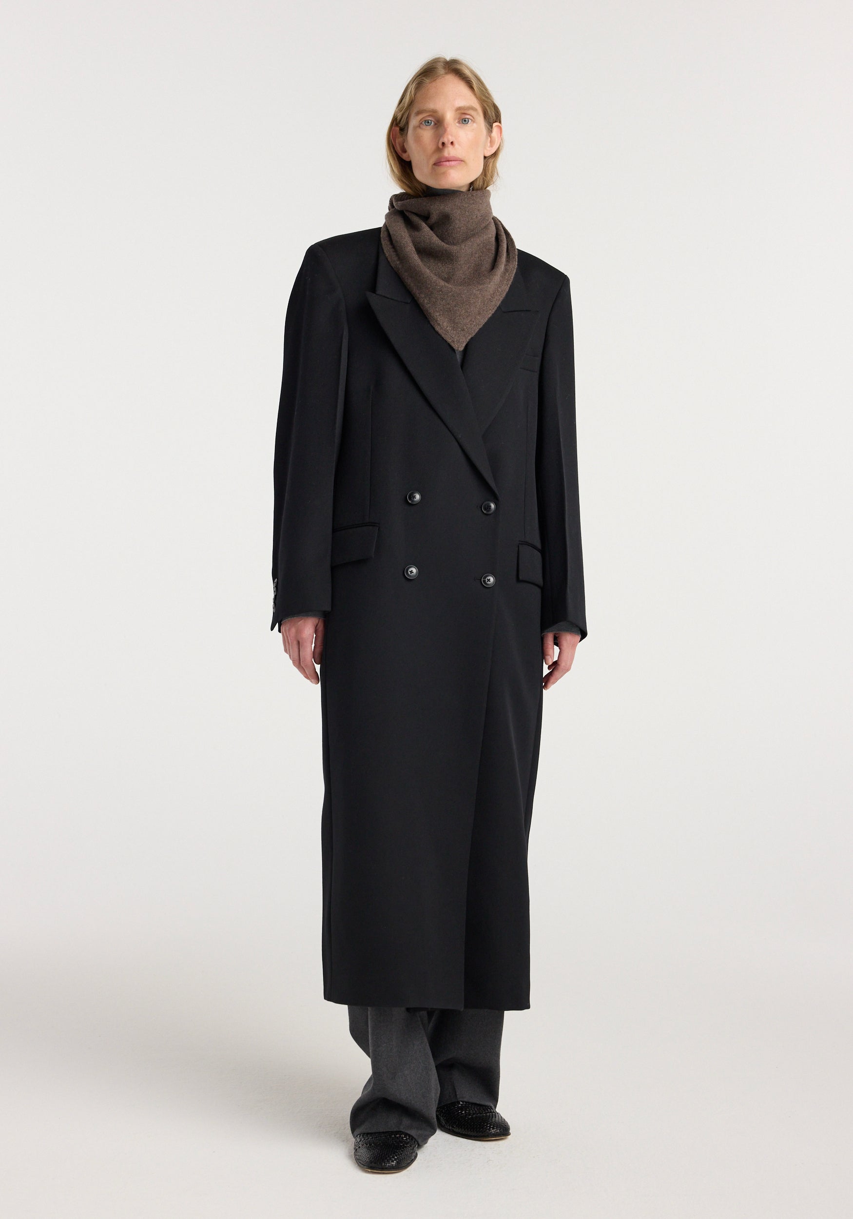 Model wearing a small knitted triangle scarf | brown melange, styled with a long black double-breasted coat and dark trousers against a plain background.