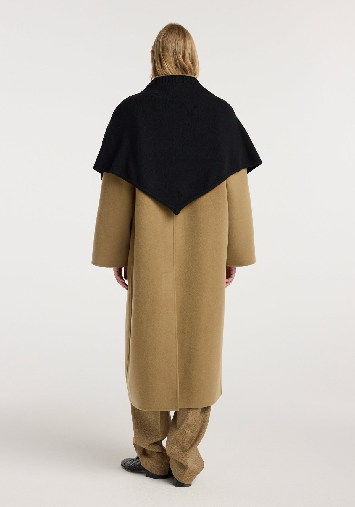Person seen from behind wearing a large knitted triangle scarf | black draped over a long beige coat against a plain light background.