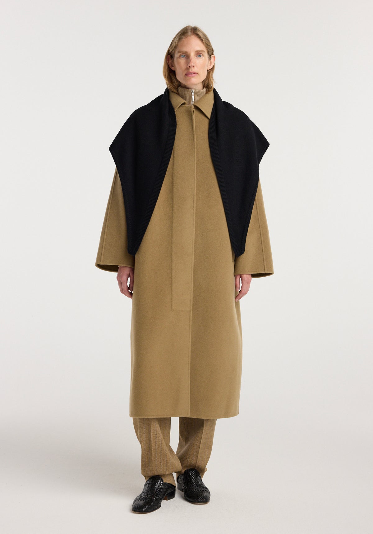 Woman wearing a beige long coat and matching pants with a Large knitted triangle scarf | black draped over her shoulders against a plain light background.