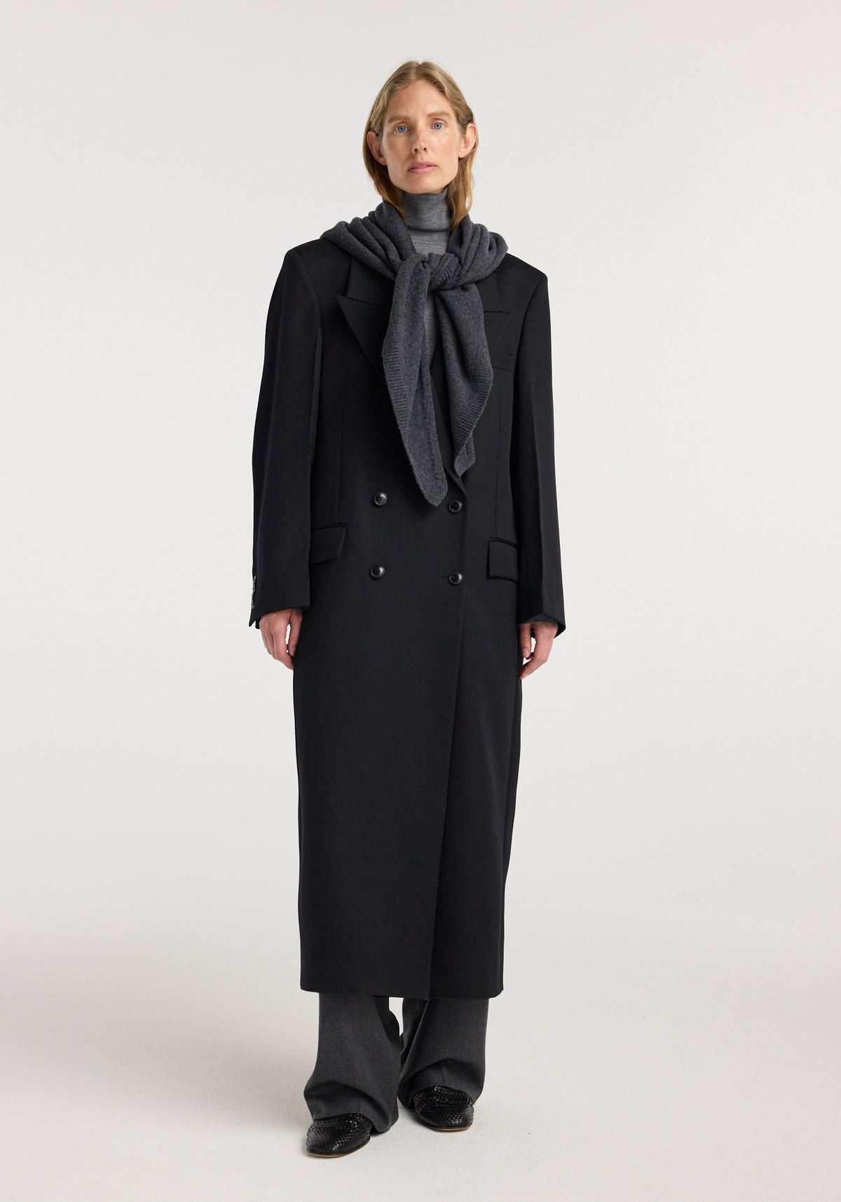 Model wearing a large knitted triangle scarf | anthracite melange, styled over a dark long coat and gray turtleneck with wide-leg pants against a plain background.