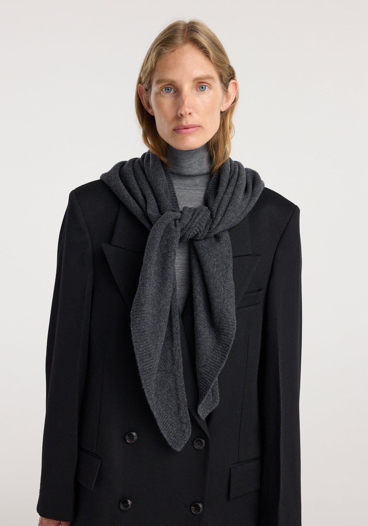 Woman wearing a large knitted triangle scarf | anthracite melange draped and knotted over a gray turtleneck and black double-breasted coat against a plain background.