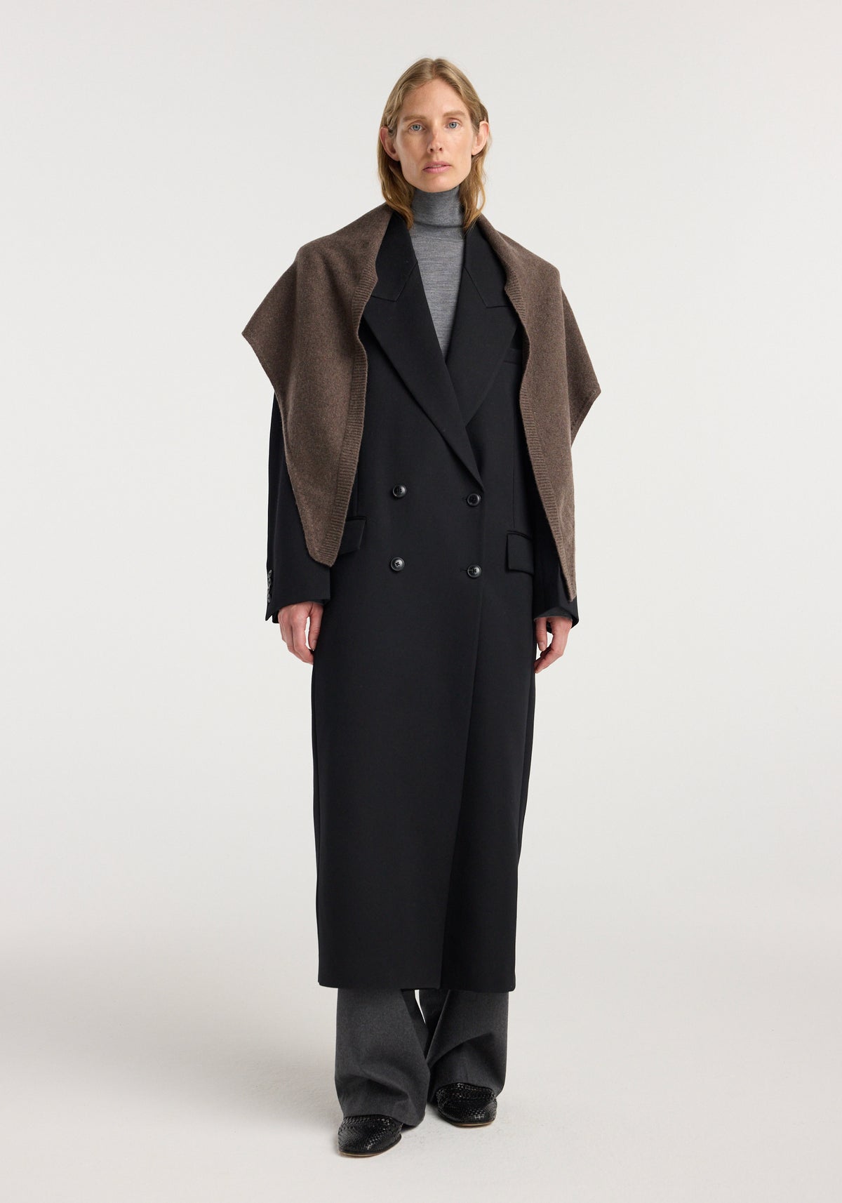 Woman wearing a Large knitted triangle scarf | brown melange draped over her shoulders, paired with a long black double-breasted coat, gray turtleneck, and gray trousers.