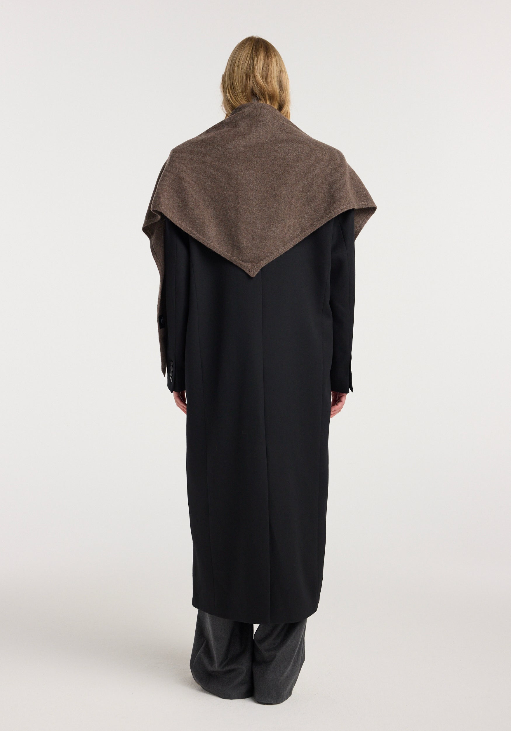 Rear view of a person wearing a Large knitted triangle scarf | brown melange draped over their shoulders paired with a long black coat and gray pants against a plain background.