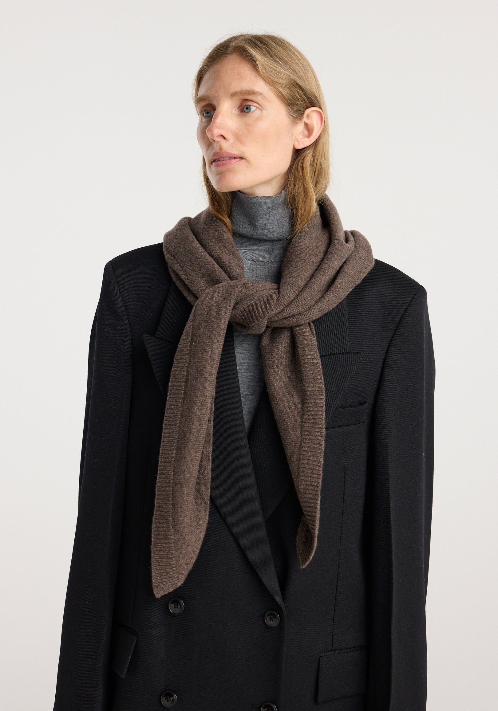 Woman wearing a large knitted triangle scarf | brown melange draped over a black double-breasted blazer and gray turtleneck sweater against a plain background.