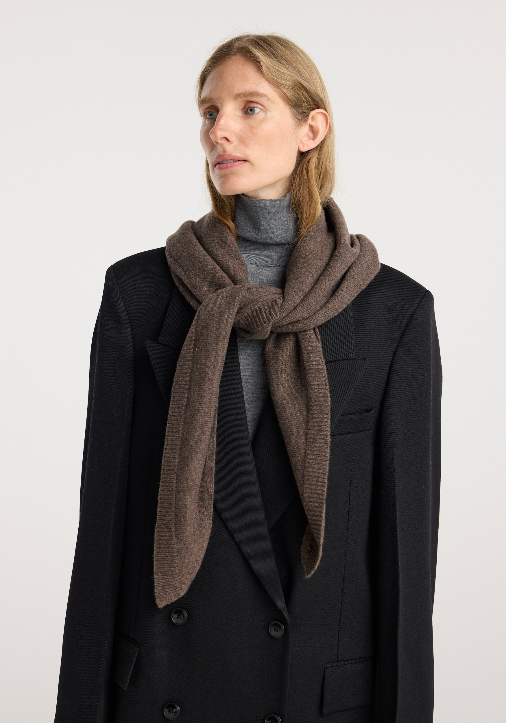 Woman wearing a large knitted triangle scarf | brown melange draped over a black double-breasted blazer and gray turtleneck sweater against a plain background.