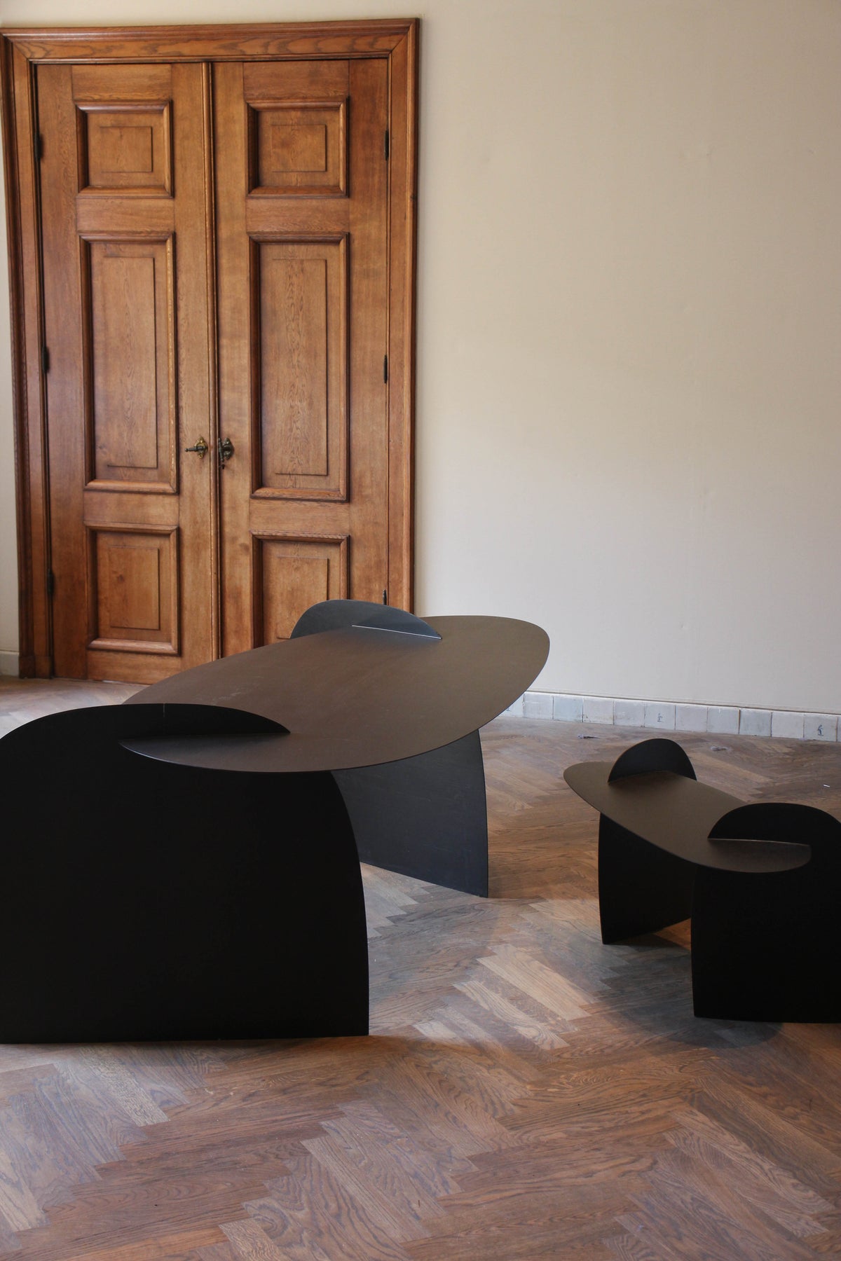 Sculpted Table | 9845: Matte-black oak table with curved, interlocking panels in a minimalist design, set in a room with herringbone wood flooring and tall doors.