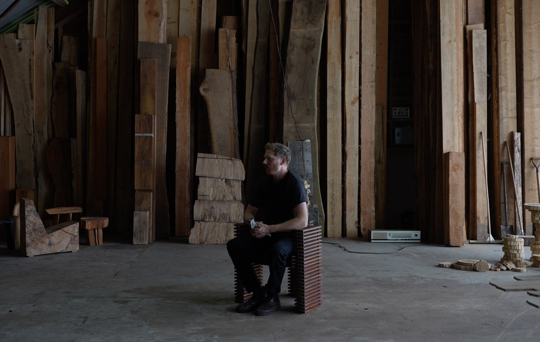 Man sitting on a chair in a room filled with wooden planks leaning against the walls 