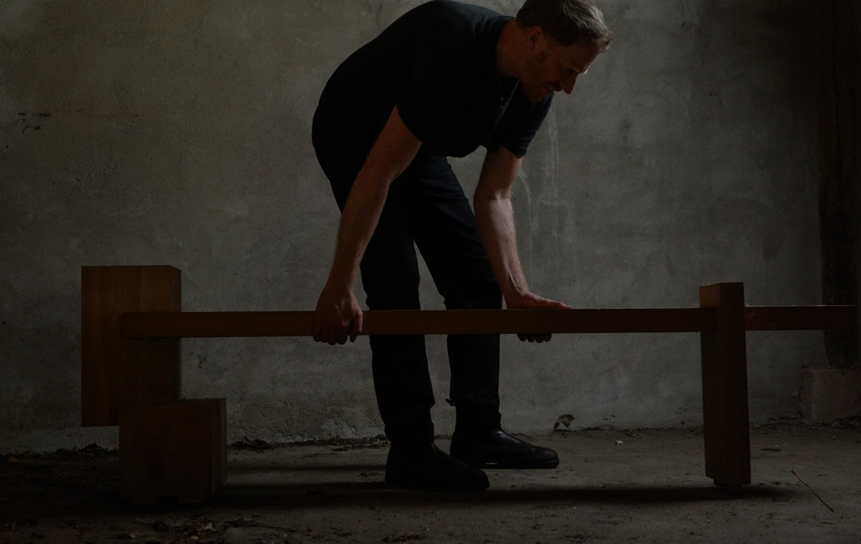 Person lifting a wooden bench in a dimly lit room with concrete walls 