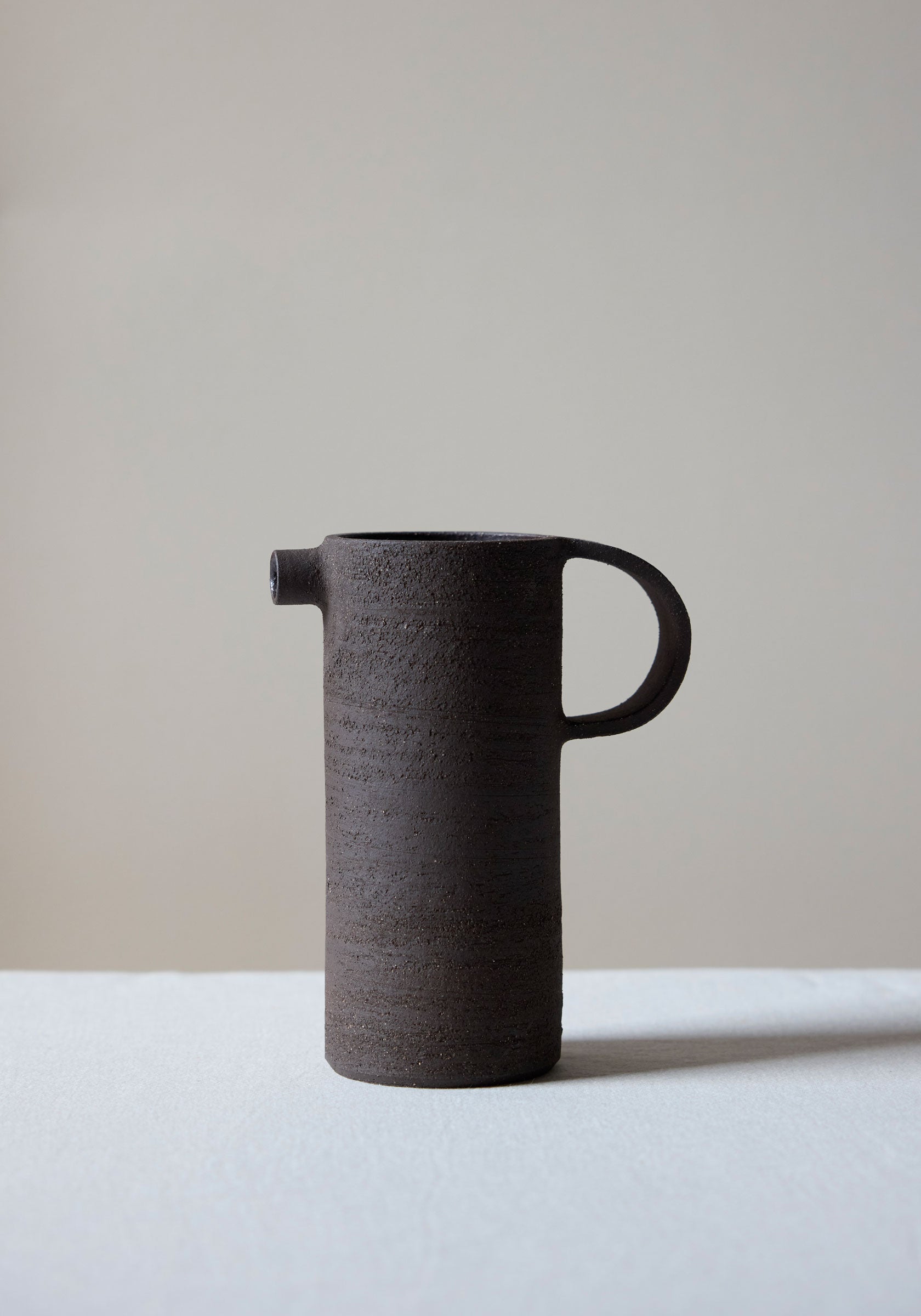 Minimalist dark brown textured ceramic carafe with a narrow spout and rounded handle standing on a light surface against a neutral background, Ceramic Carafe | soil.
