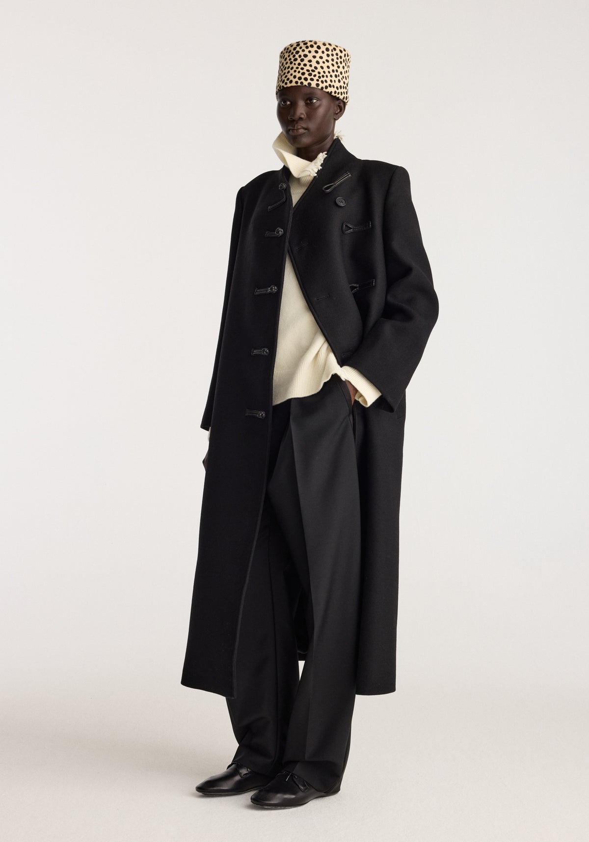 Cheetah leather hat | cheetah worn with a black long coat, cream sweater, wide trousers, and black shoes on a model in a studio setting
