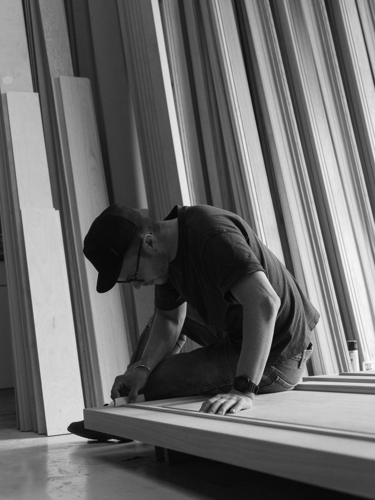Black and white photo of a worker wearing a cap kneeling beside a large window frame and using a hand tool while working on the structure