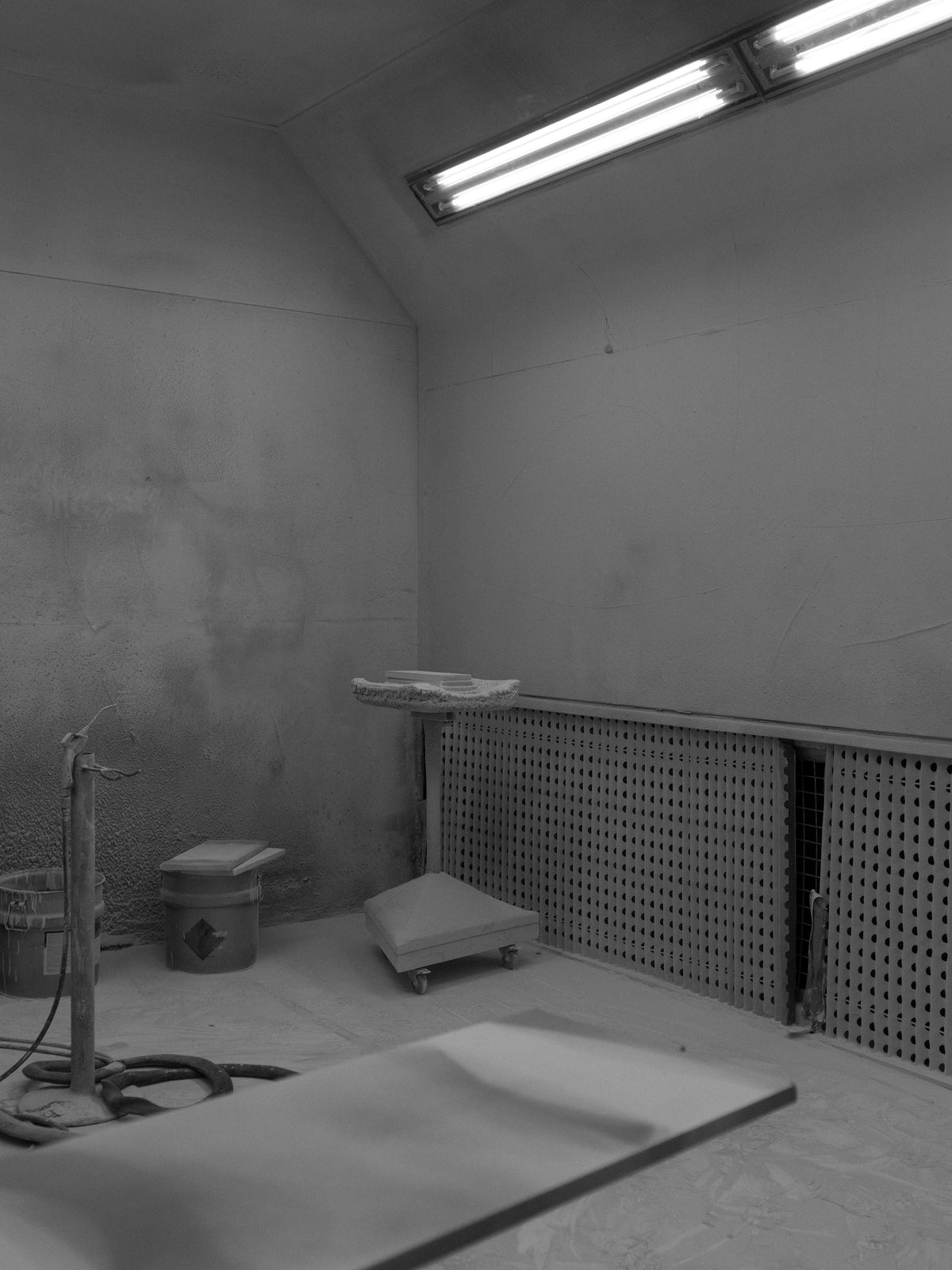 Empty industrial workshop with fluorescent ceiling lights, gray walls, perforated metal panels, a worktable, wheeled equipment, and coiled hoses on the floor.