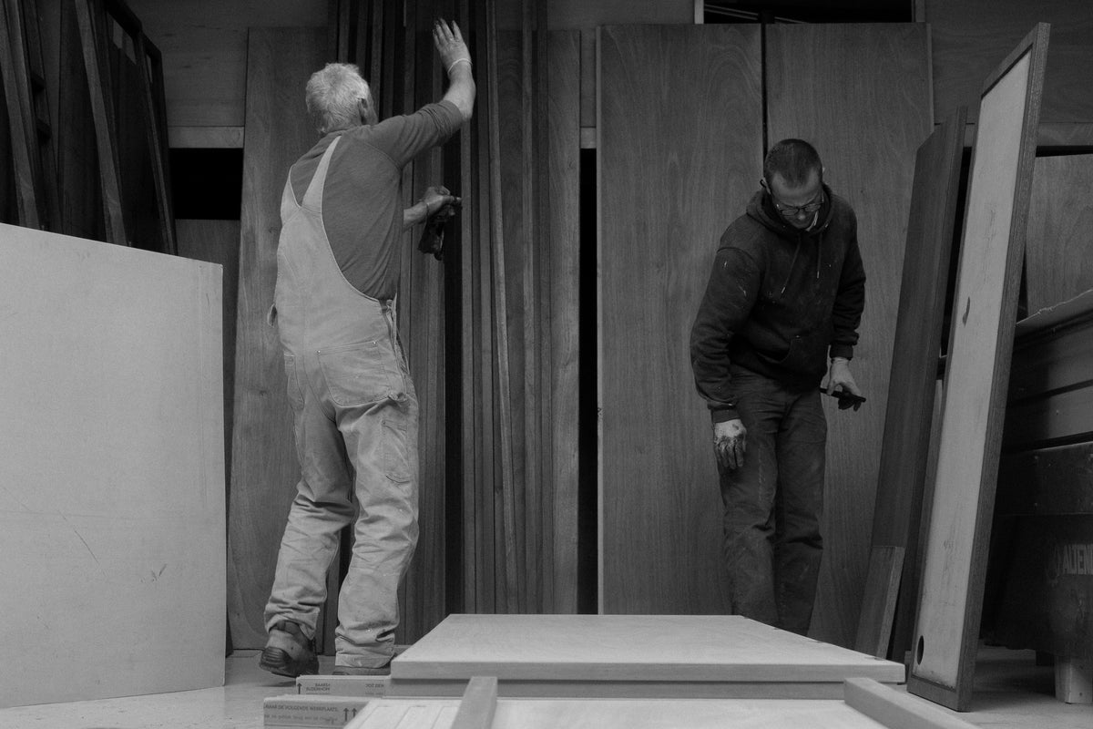Black and white photo of two workers in a workshop, with one reaching toward a wall while the other inspects materials near a large workbench.