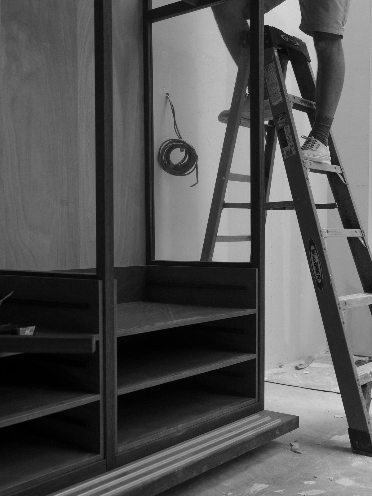 Black and white photo of a worker on a step ladder beside a partially assembled dark shelving unit with open compartments and a coiled cable on the wall