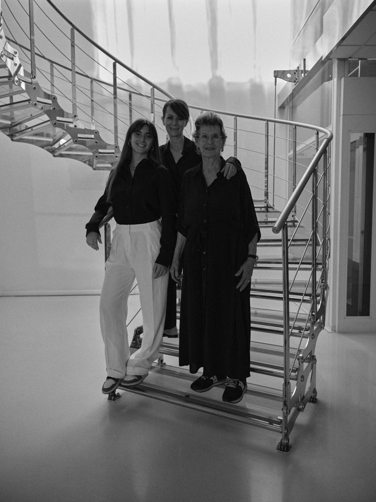Three generations of women, grandmother, mother and daughter, standing on a modern staircase with glass railings.