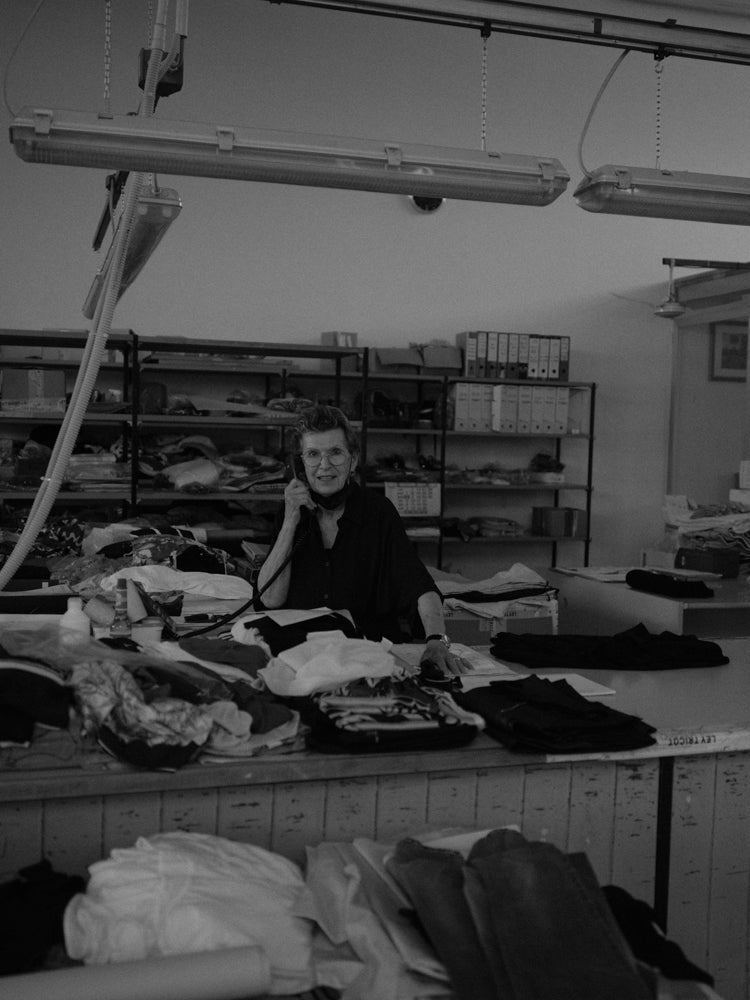 Woman sitting at a table with various fabrics and clothes folded on top, holding a cord telephone in her hand in a room with shelves in the background