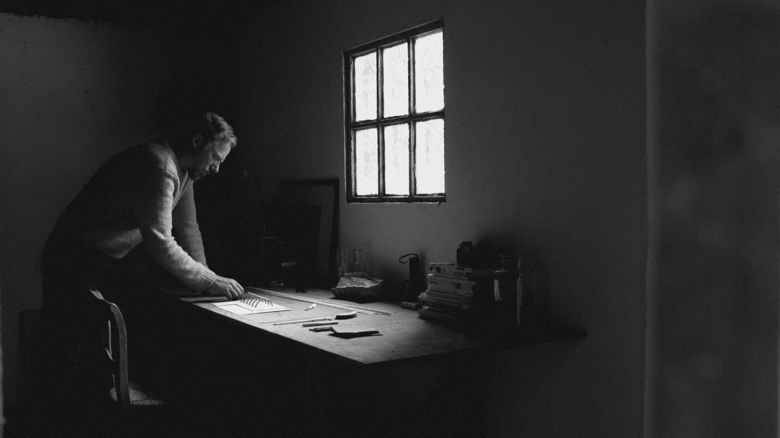 Black and white image of a man standing next to a desk in a room with a small window with light filtering through