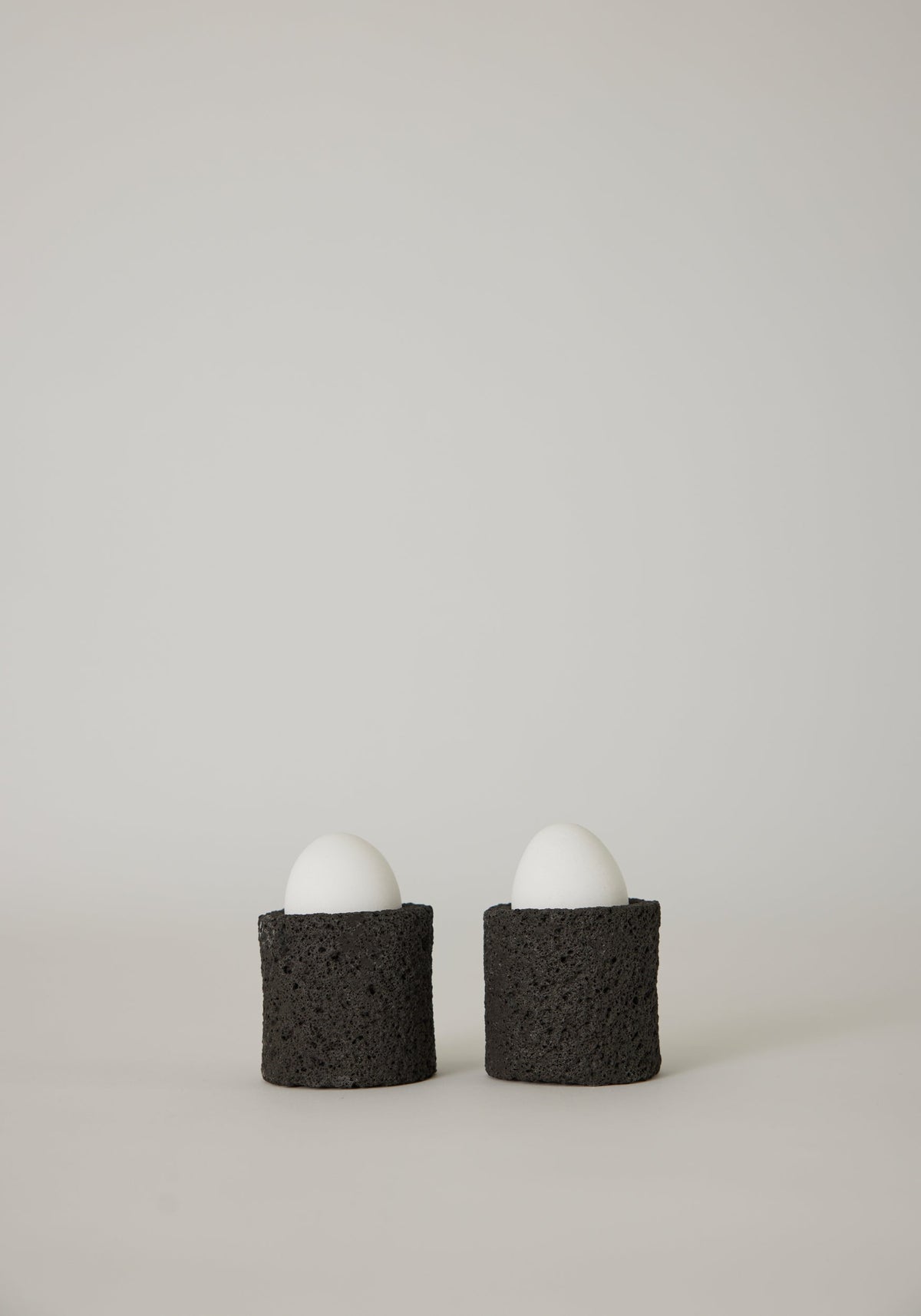 Egg Cups: Two white eggs in handcrafted black holders made from lava stone, showcasing a unique porous texture, designed by artist Louis Crombag.